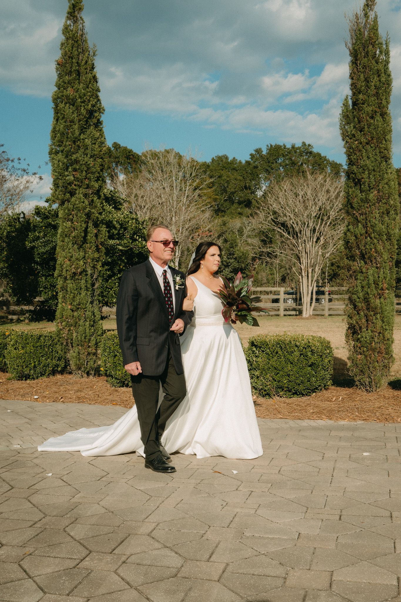 Bride and groom walk together on a stone path flanked by tall cypress trees at an outdoor wedding venue.