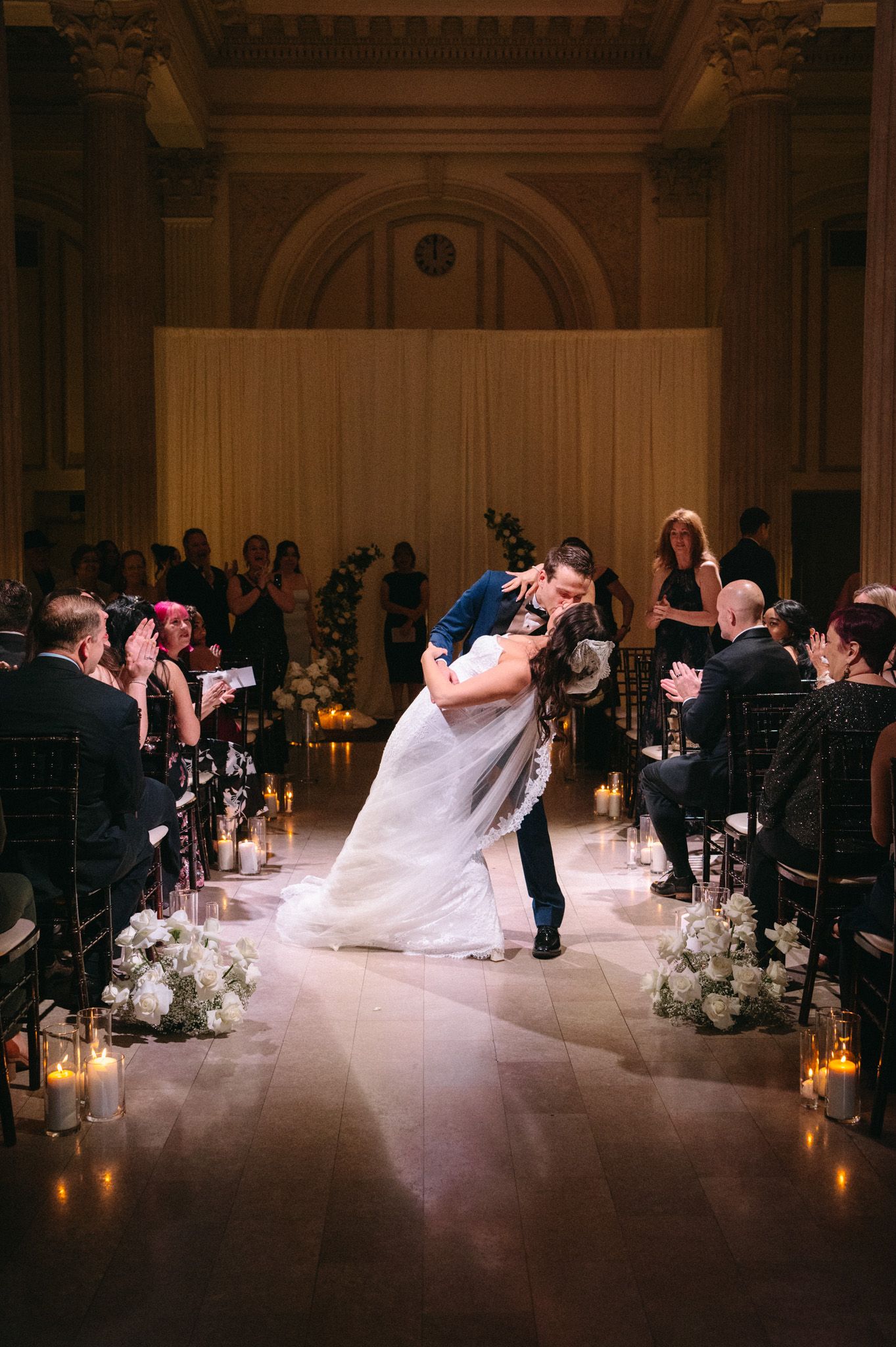 Bride and groom share a romantic dip kiss at wedding ceremony aisle with candles and white floral arrangements.