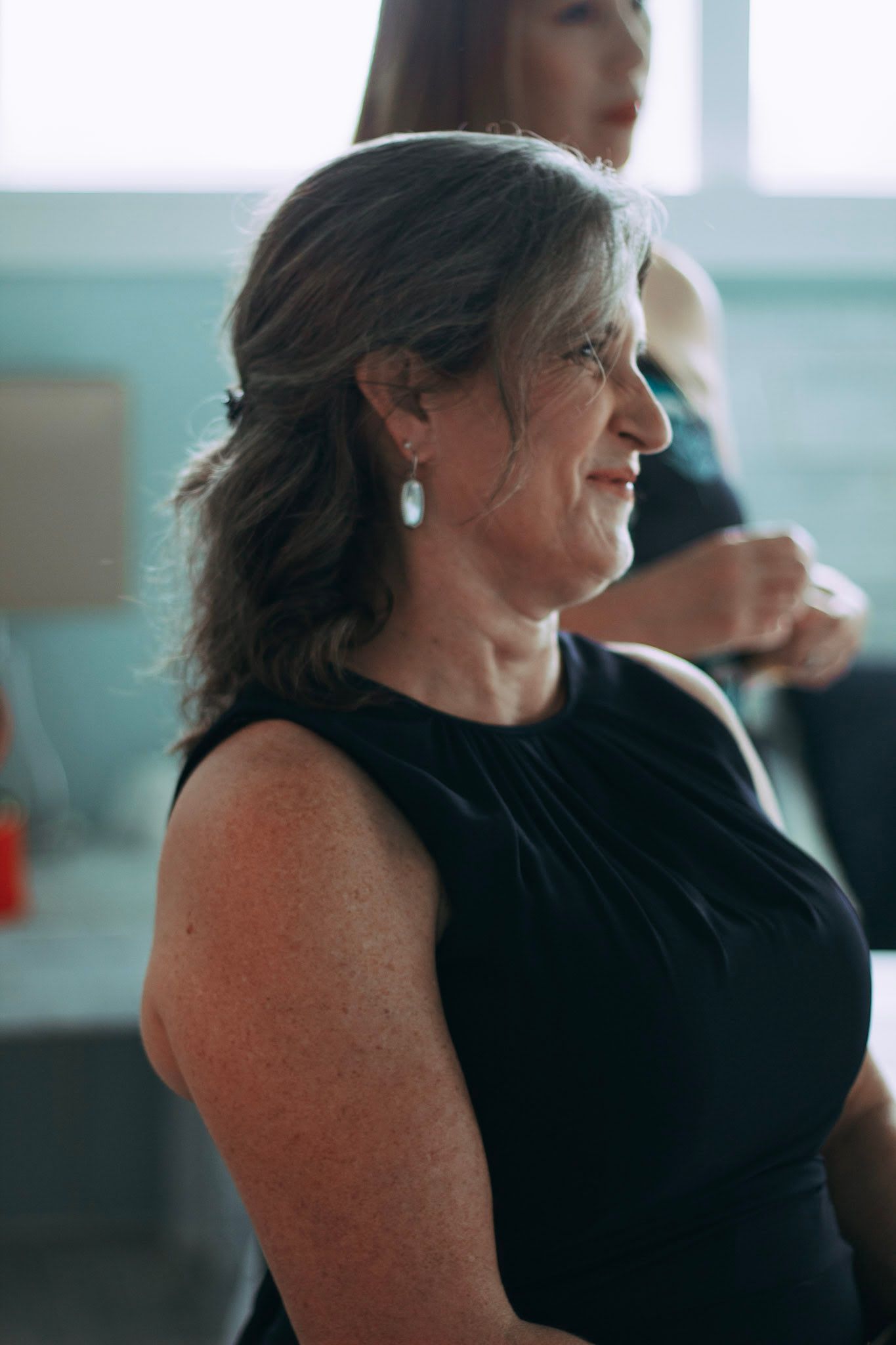 Elegant older woman in black sleeveless dress with drop earrings at wedding event.