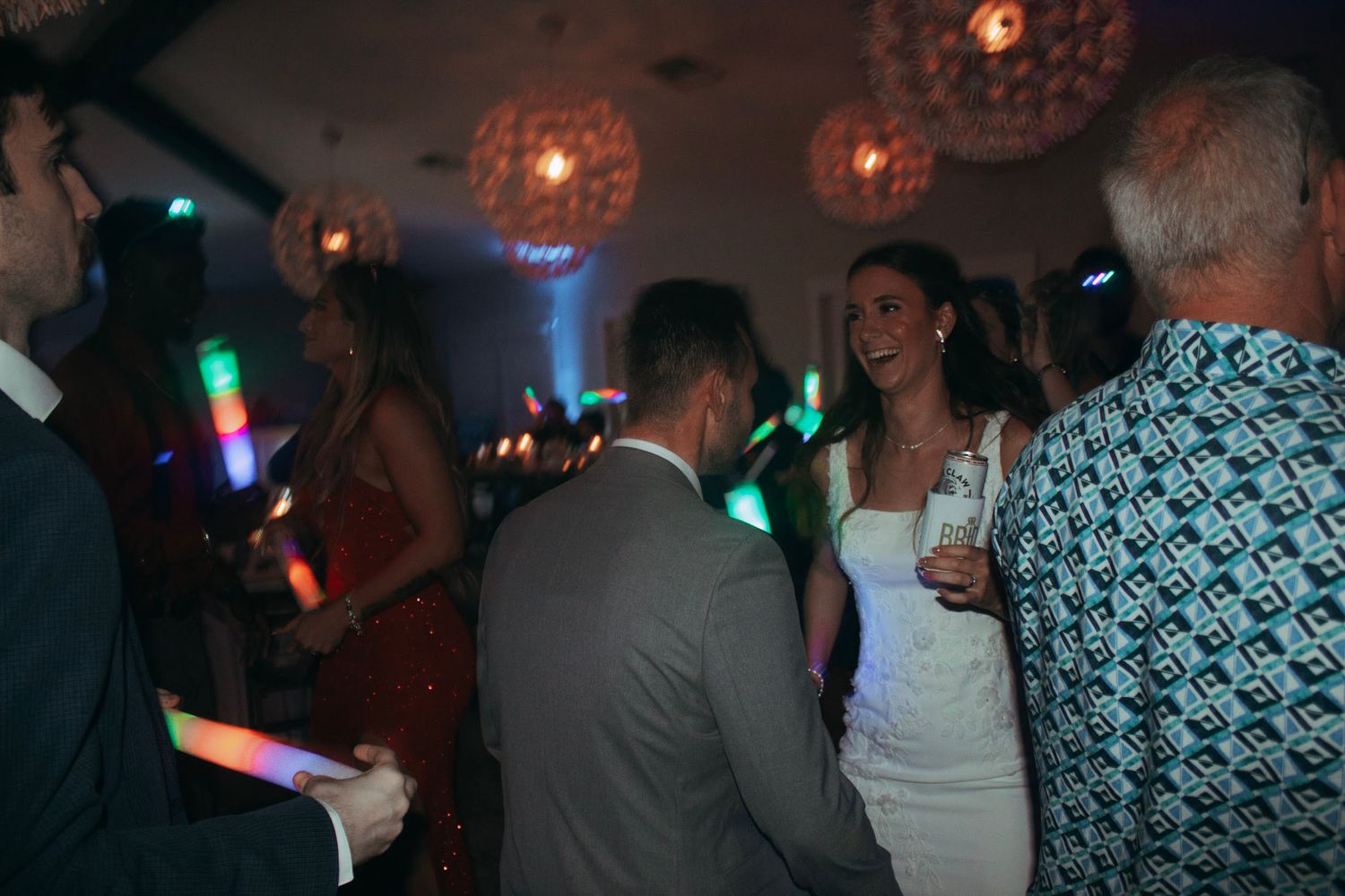 Bride and groom dancing with guests at colorful wedding reception with glowing light decorations.