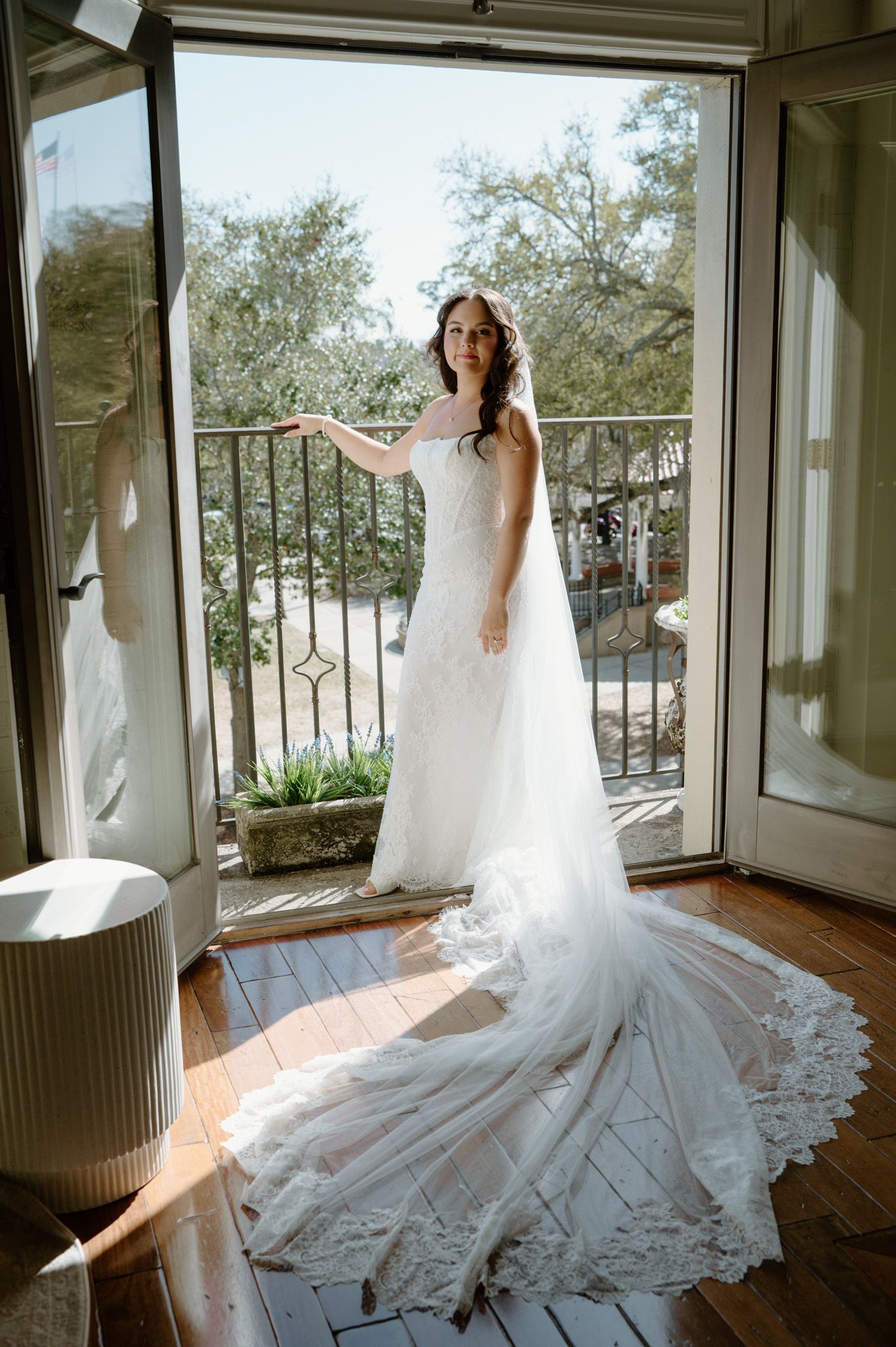 Bride in a strapless lace wedding gown with long train stands on a sunlit balcony doorway.