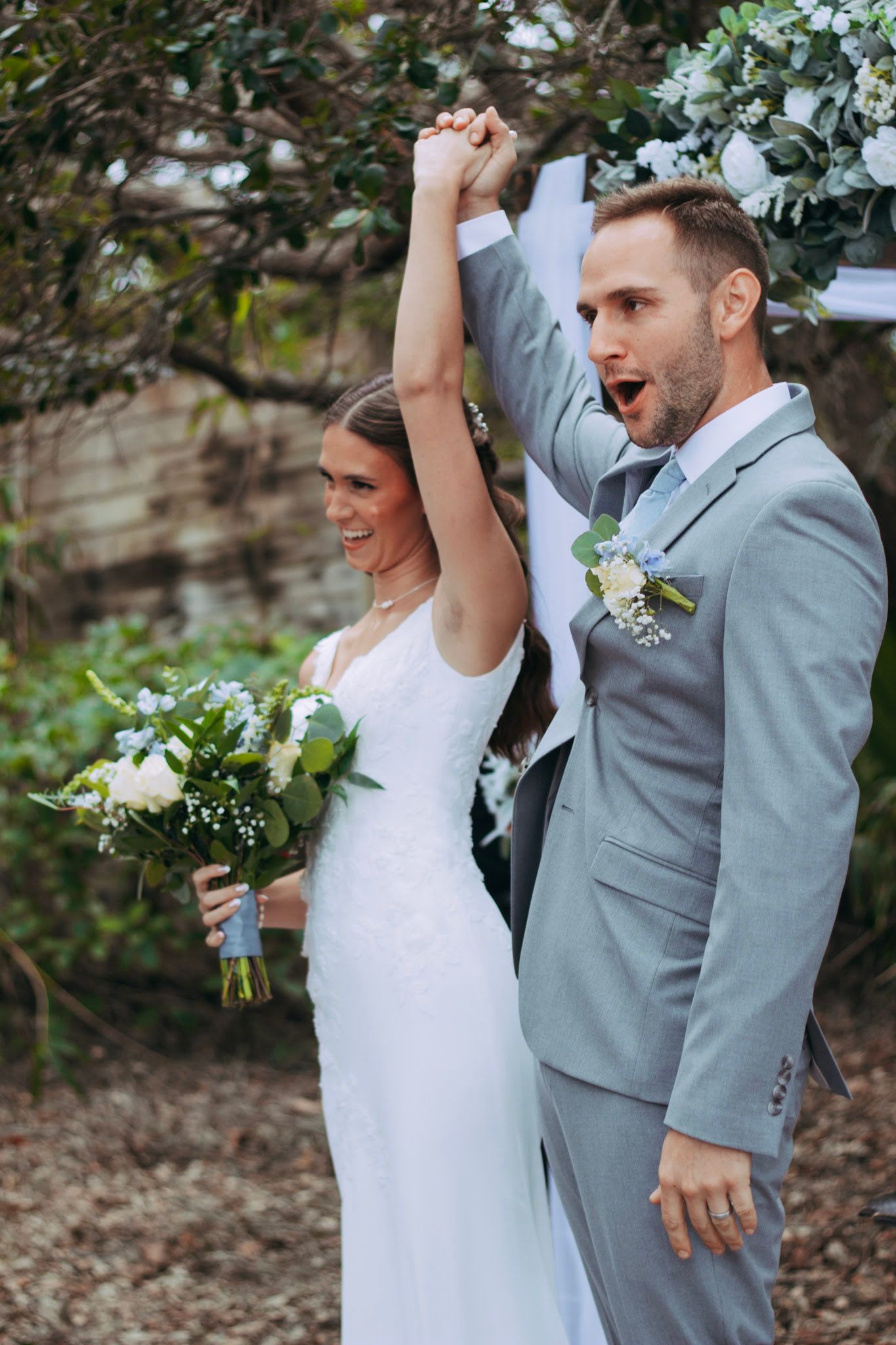 Excited newlyweds raise hands in celebration holding bouquet after outdoor garden wedding ceremony.
