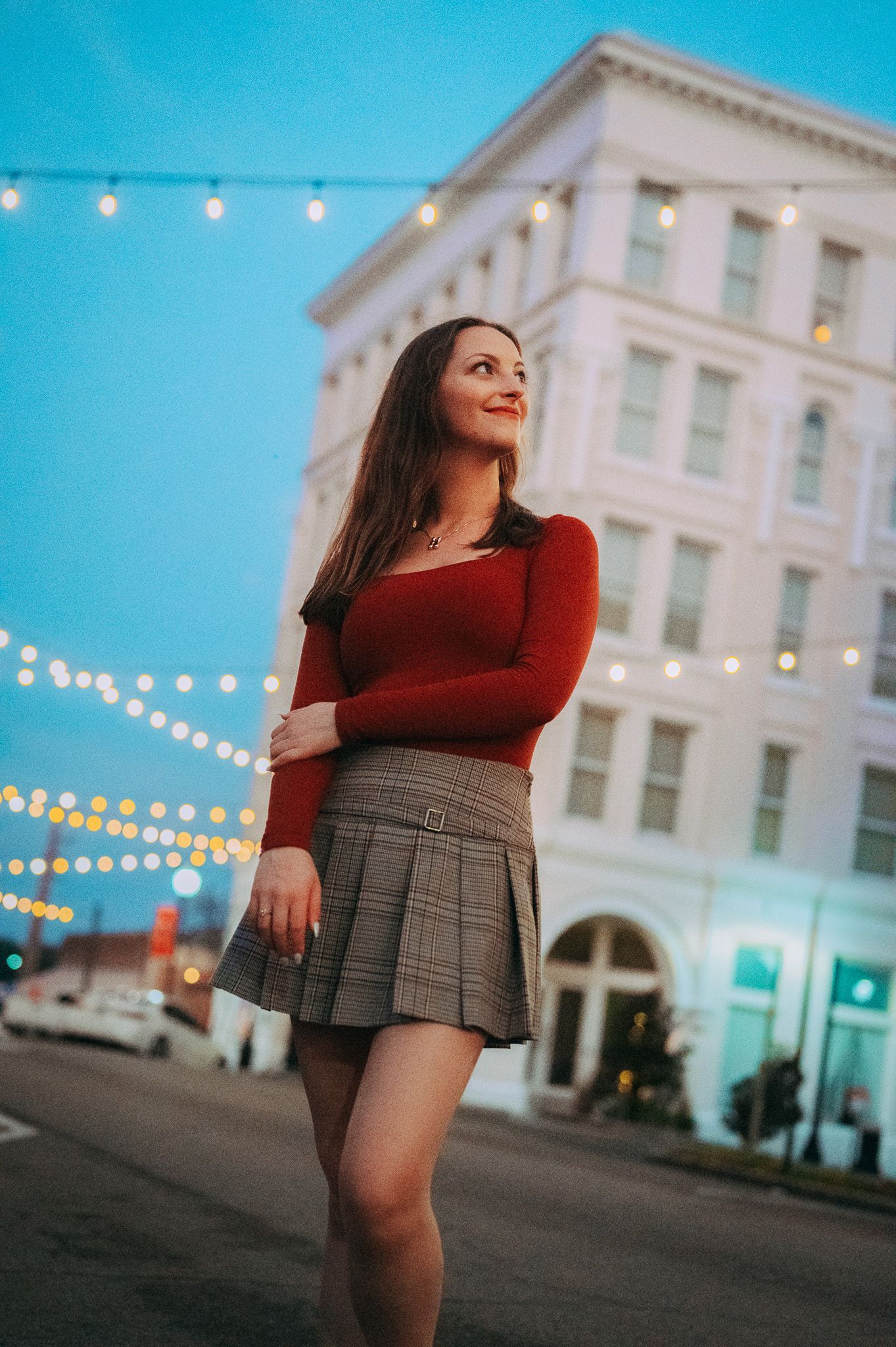 Young woman in red top and plaid skirt smiling under string lights at dusk.