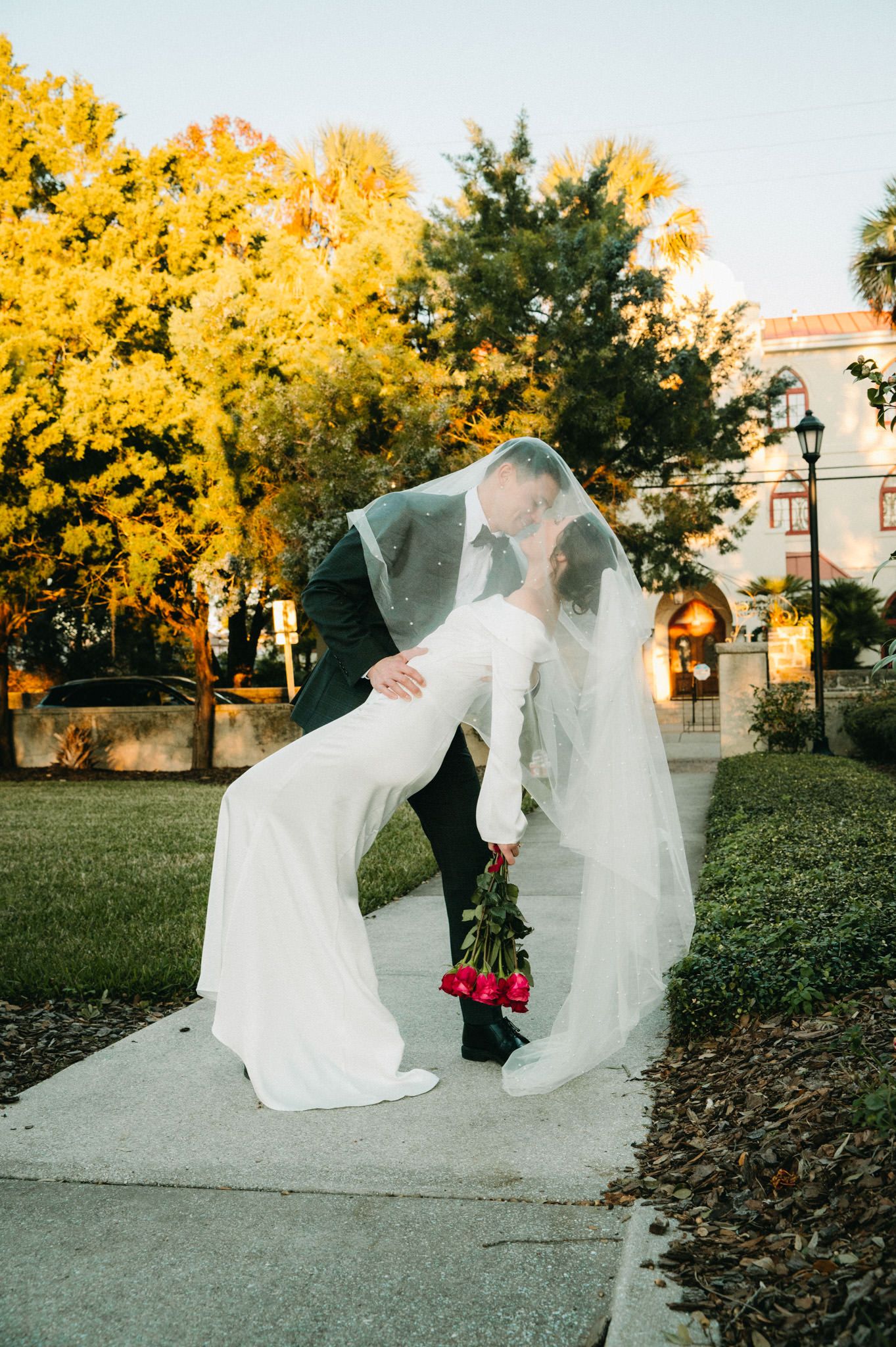 Bride and groom share a dip kiss under a veil holding red roses amid golden autumn trees.