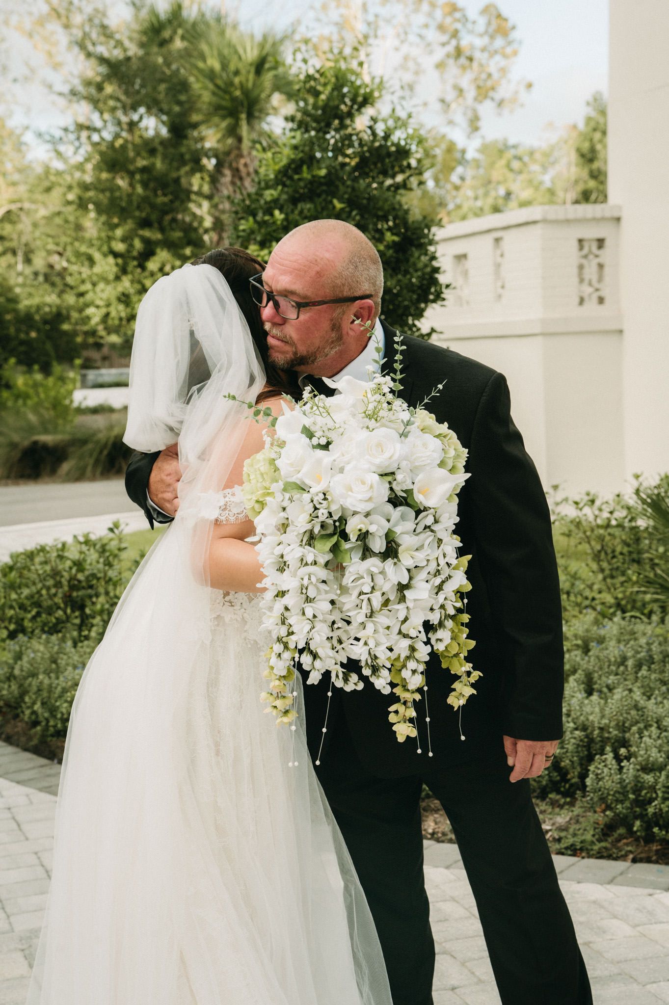 Father of the bride shares a tender kiss with bride holding a stunning white cascading floral bouquet.