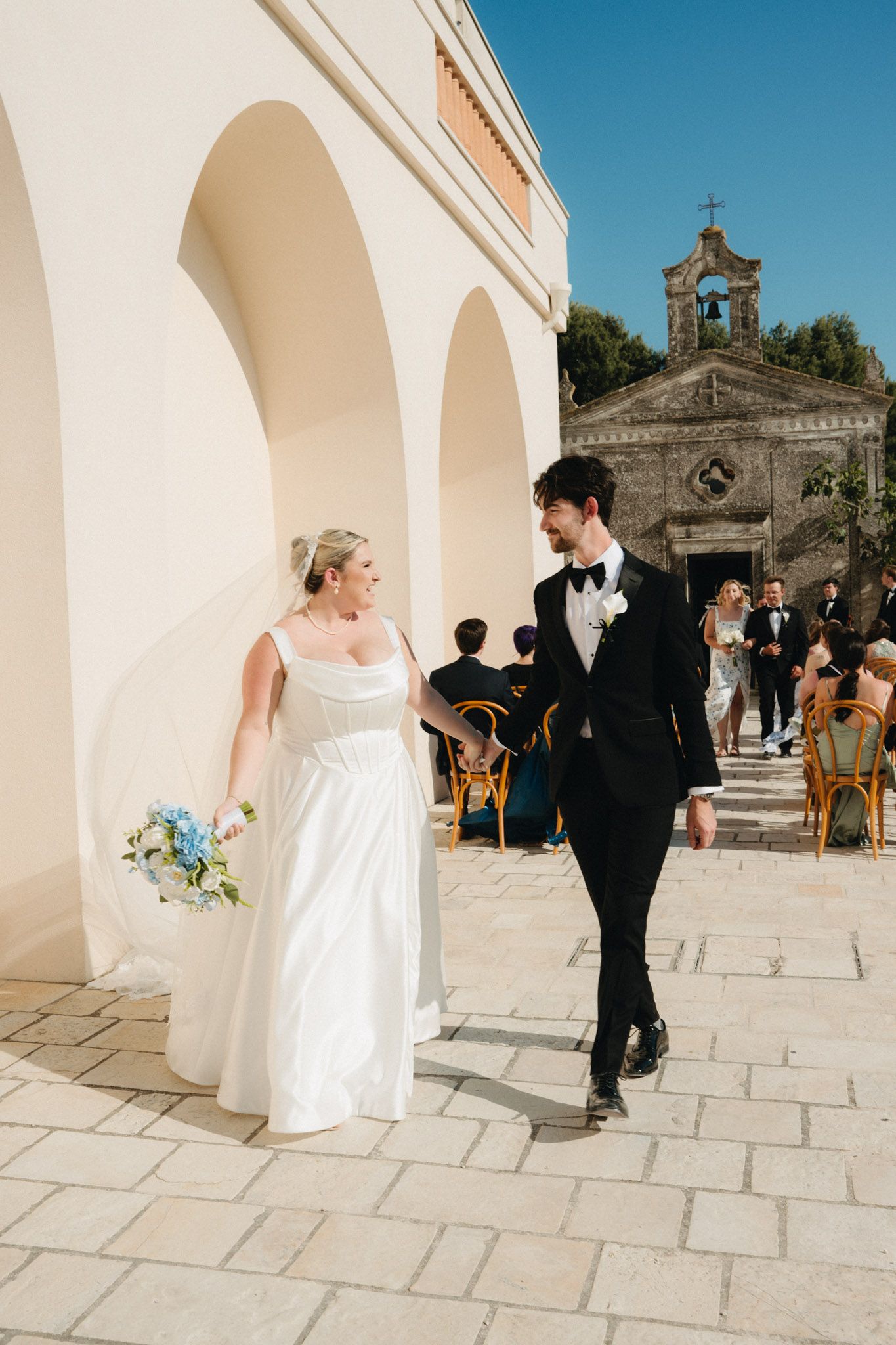 Bride and groom walk together outside a sunlit Italian church, she holds blue flowers, he wears a black tuxedo.