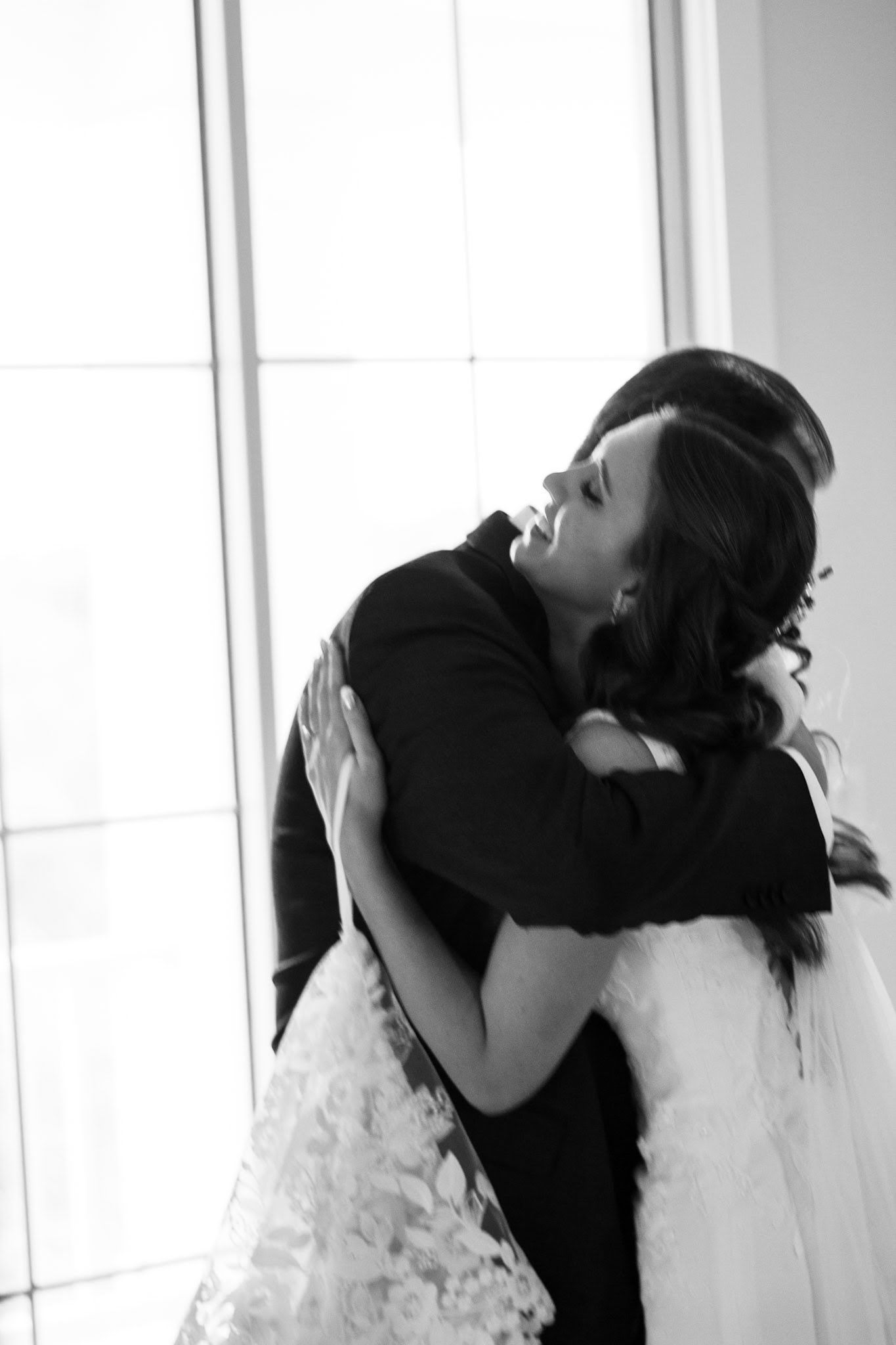 Black and white photo of bride and groom sharing a heartfelt embrace near bright window.