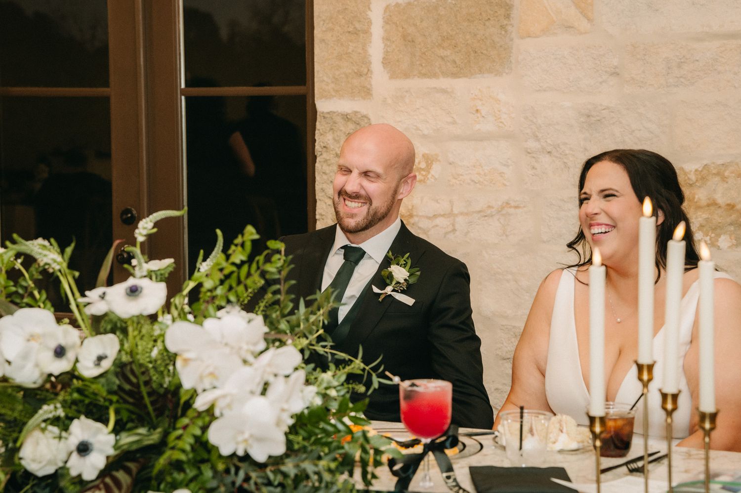 Bride and groom laughing joyfully at sweetheart table with white floral centerpiece during wedding reception.