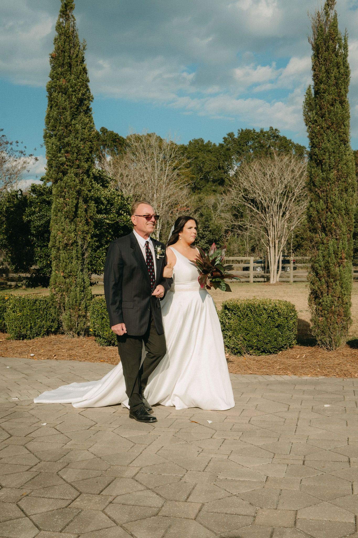 Bride walks with escort past cypress trees at outdoor wedding venue.