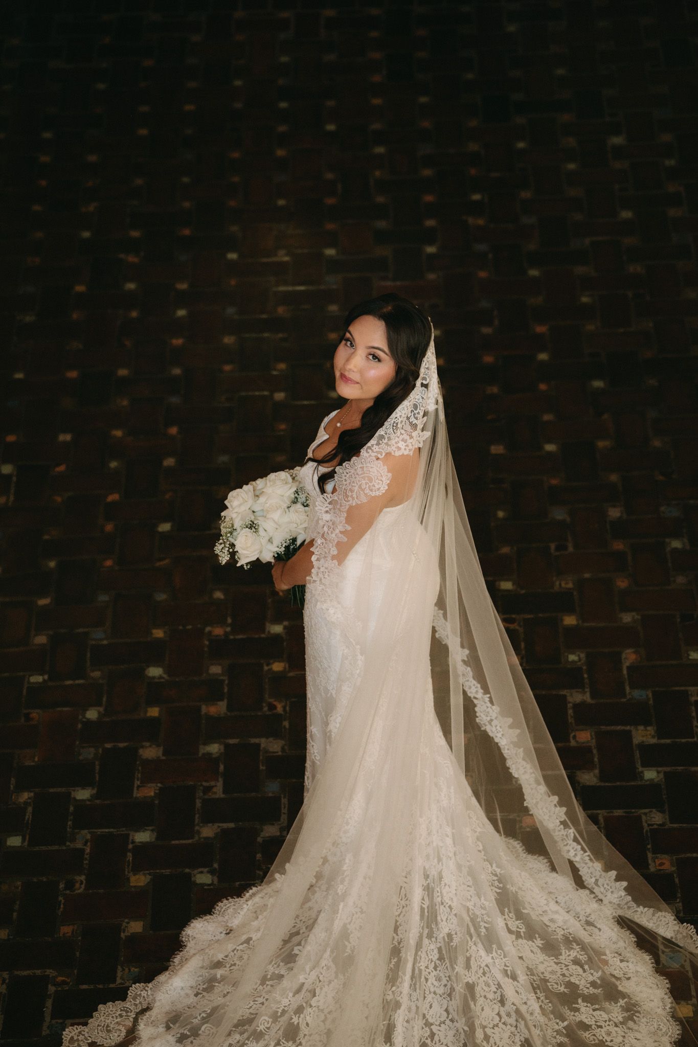 Elegant bride in lace mermaid wedding gown with cathedral veil holding white bouquet against dark brick wall.