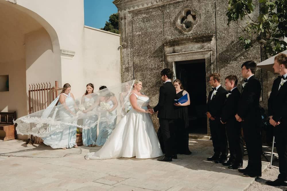 Outdoor wedding ceremony with bride in white gown, bridesmaids in blue, and groomsmen in black suits by a stone church.