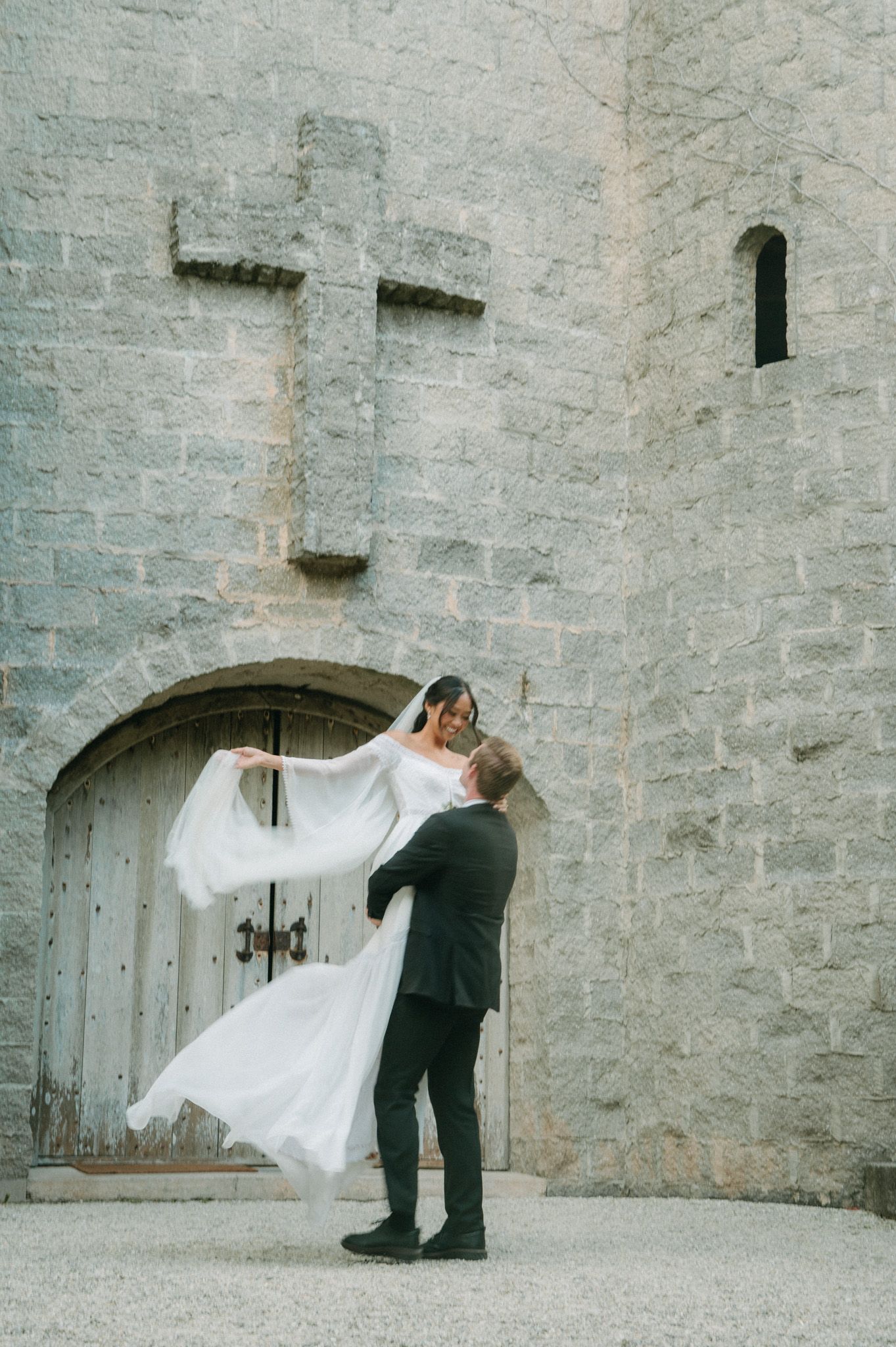 Groom lifts smiling bride in white gown before a stone wall with a large cross carved above an arched doorway.