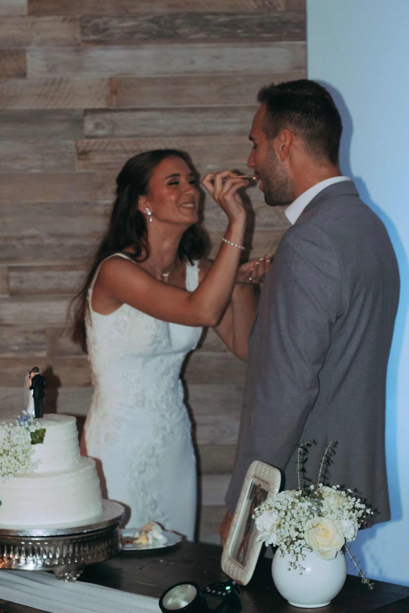 Bride in white lace dress feeding cake to groom at wedding reception with two-tier white cake.