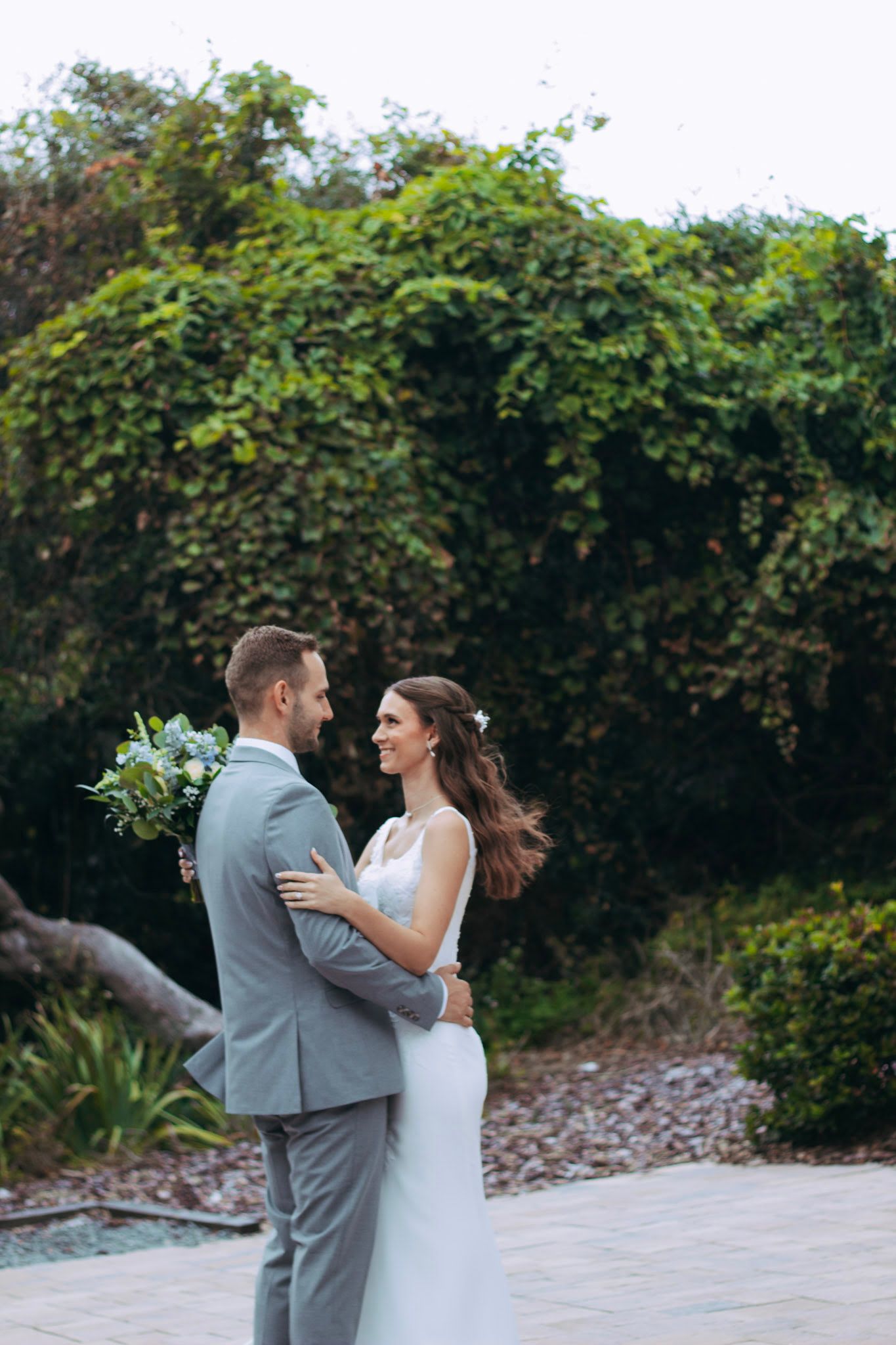 Smiling bride and groom embracing outdoors with green ivy wall backdrop on wedding day.