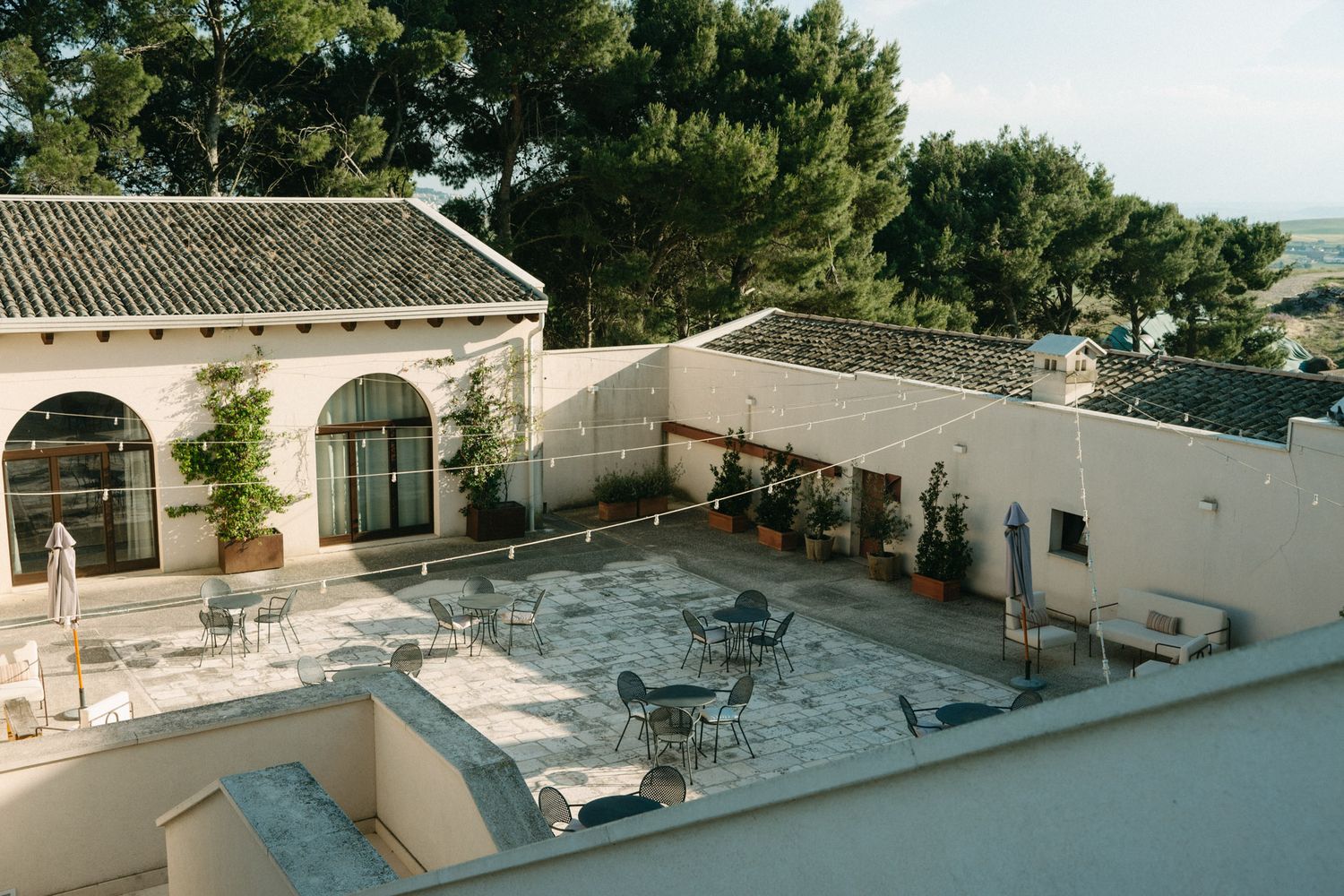 Aerial view of a Mediterranean-style courtyard with outdoor seating, white walls, tiled roofs, and lush green trees.