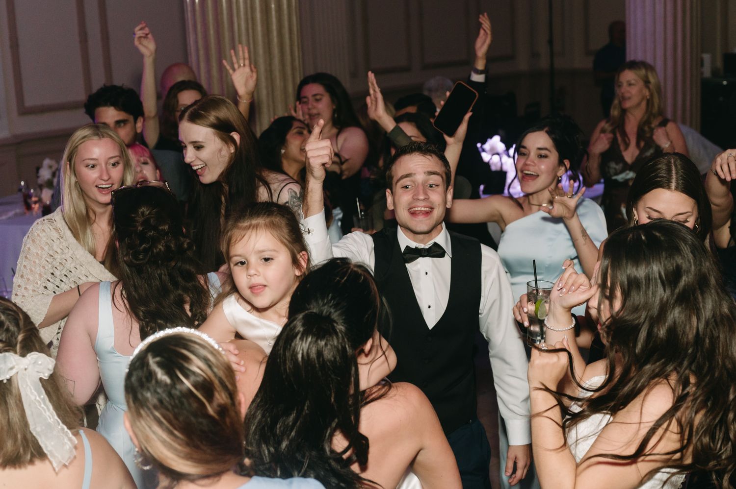 Young boy in tuxedo celebrates at formal party with crowd of guests dancing and raising hands in elegant ballroom.