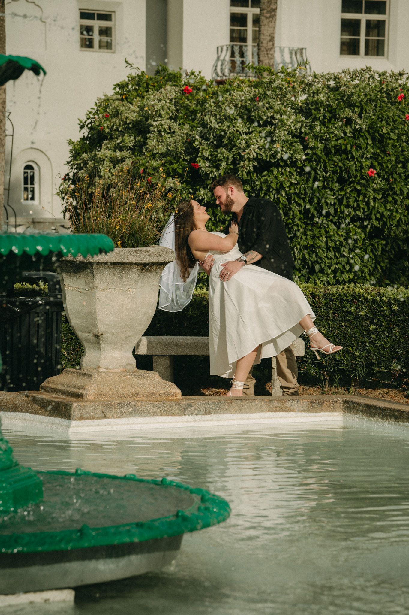 Bride and groom sharing romantic kiss seated on stone bench beside ornate garden fountain.