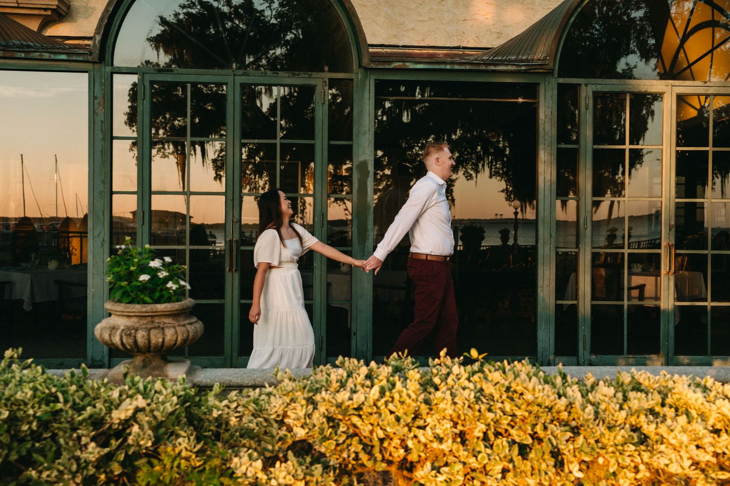 Couple holds hands walking past golden flowers at sunset near elegant glass greenhouse doors.