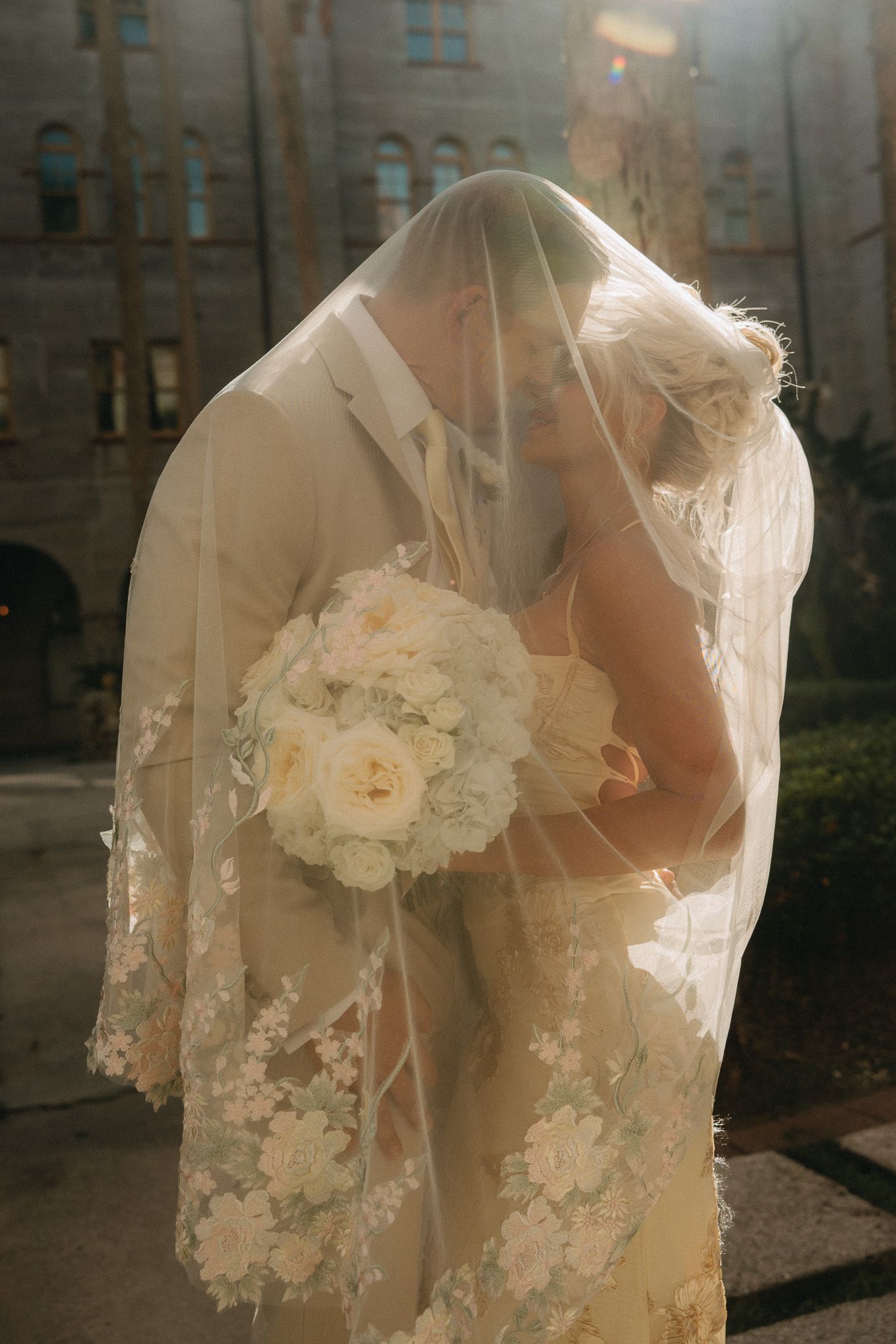 Bride and groom kissing under lace veil, bride holding white rose bouquet, romantic wedding portrait outdoors.