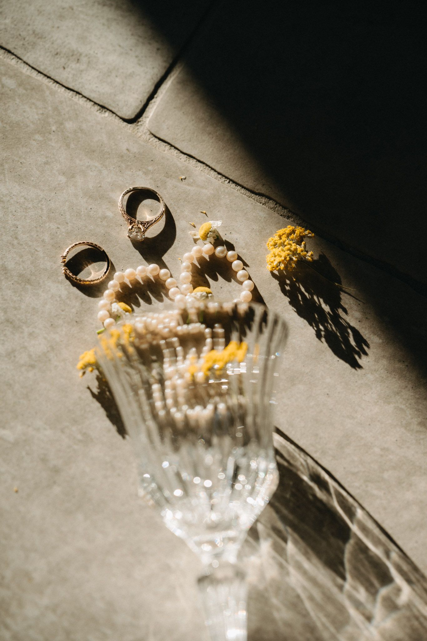 Crystal vase with pearl bracelet and yellow marigolds beside two gold rings on concrete surface in sunlight.