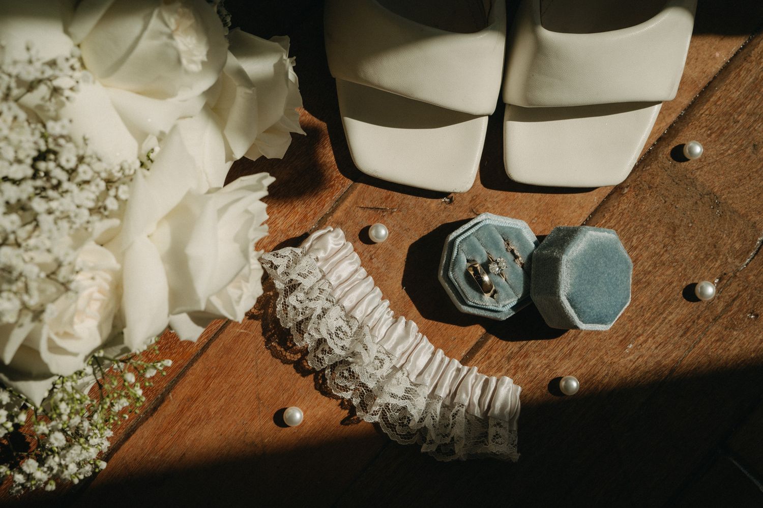 Wedding flat lay with lace garter, velvet ring box, pearl pins, white roses, and bridal heels on wooden surface.