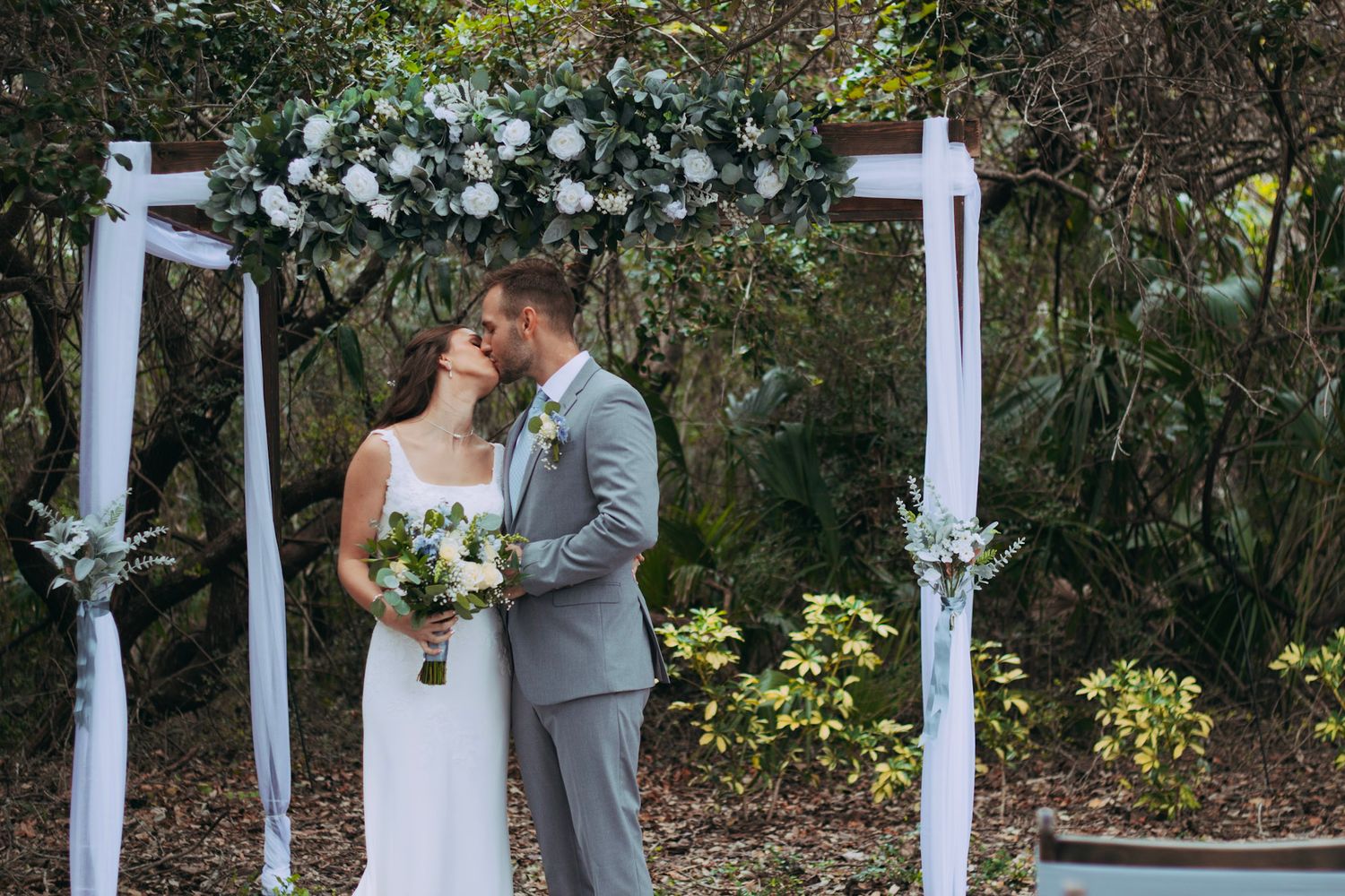 Bride and groom sharing first kiss under floral wedding arch decorated with white roses.