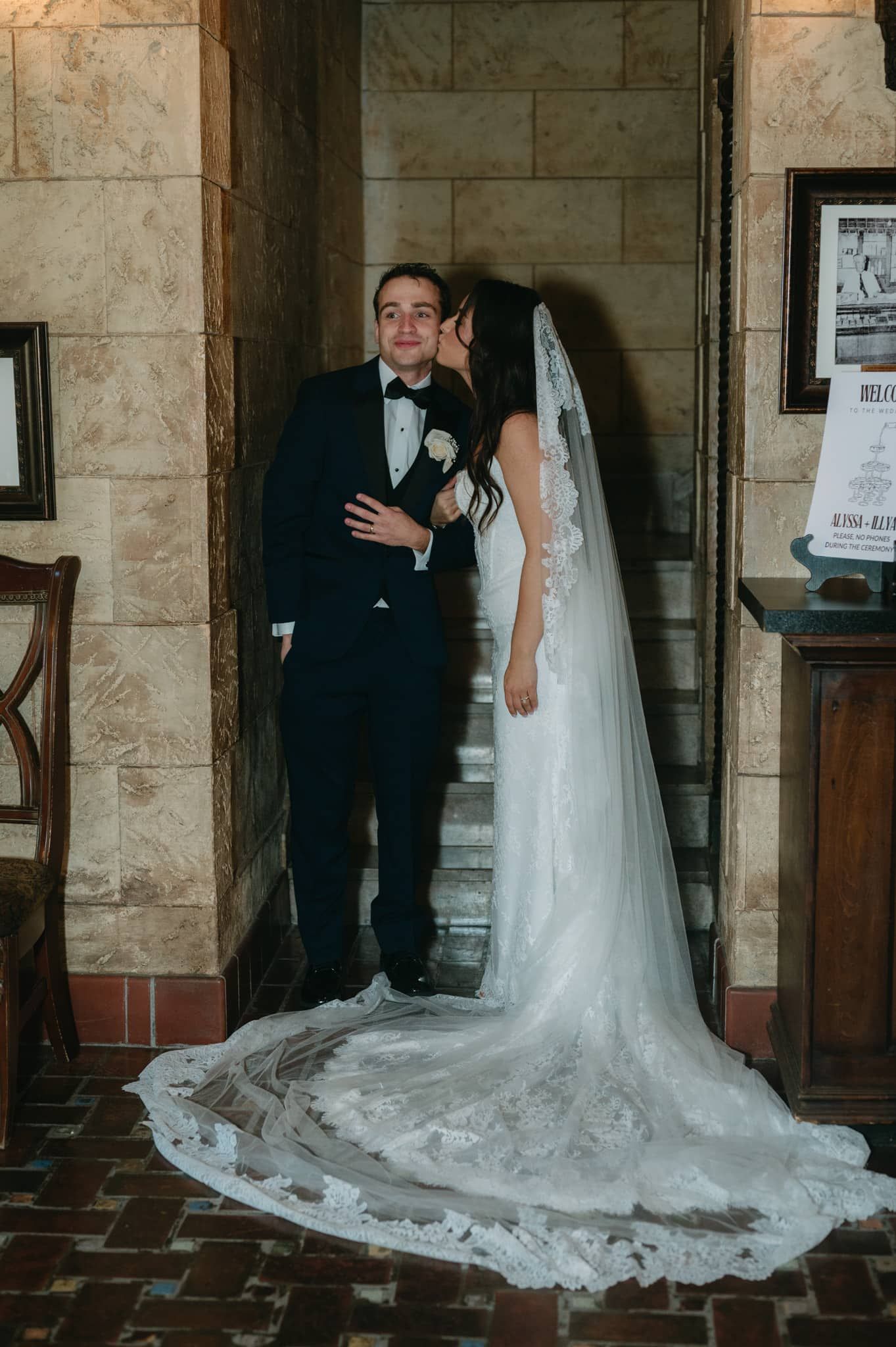 Bride in lace gown and veil kisses groom in navy tuxedo on brick staircase at The Treasury on the Plaza in St. Augustine, FL.
