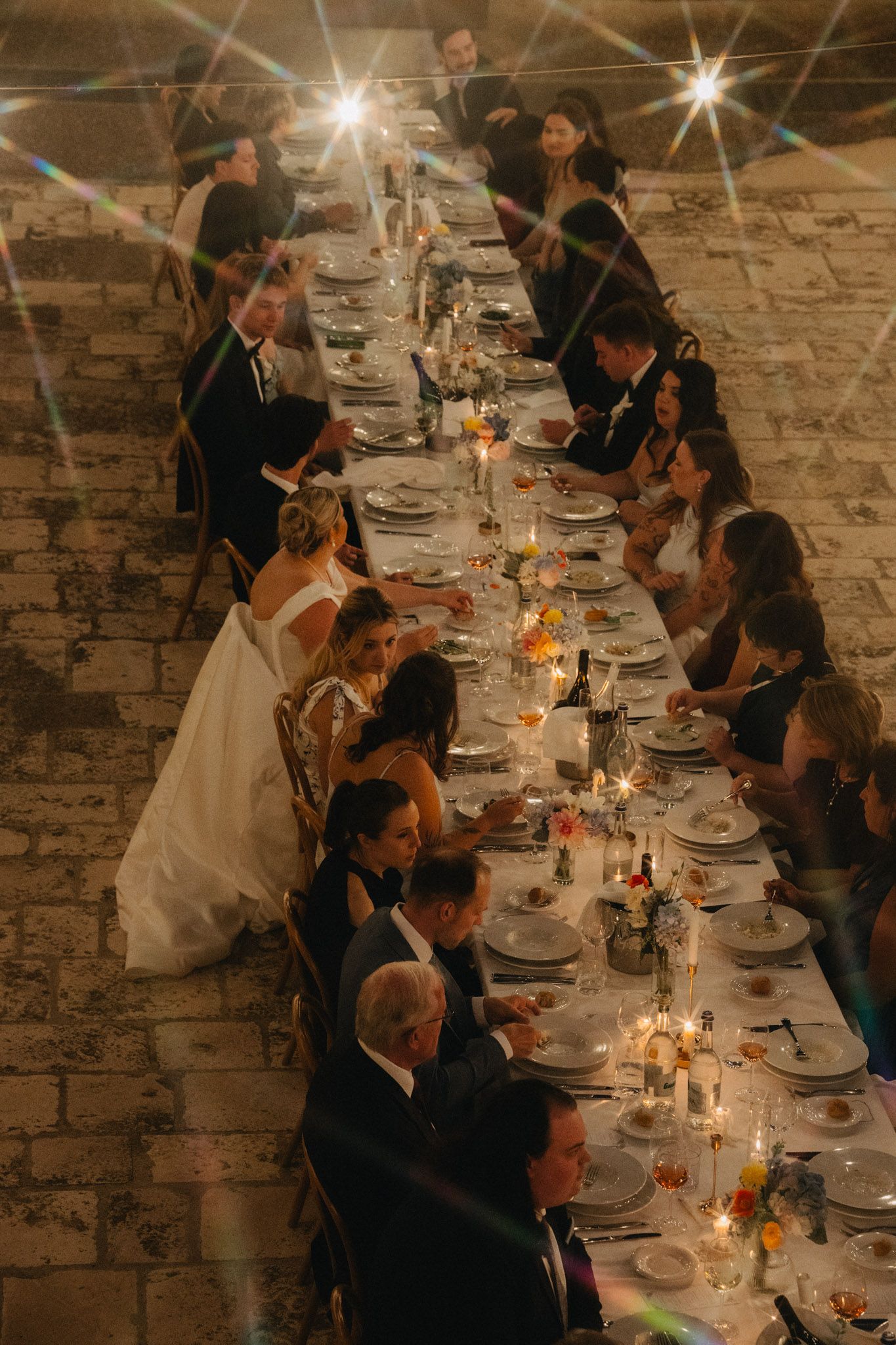 Aerial view of wedding reception dinner at long candlelit table with bride in white gown, guests in formal attire, stone wall venue.