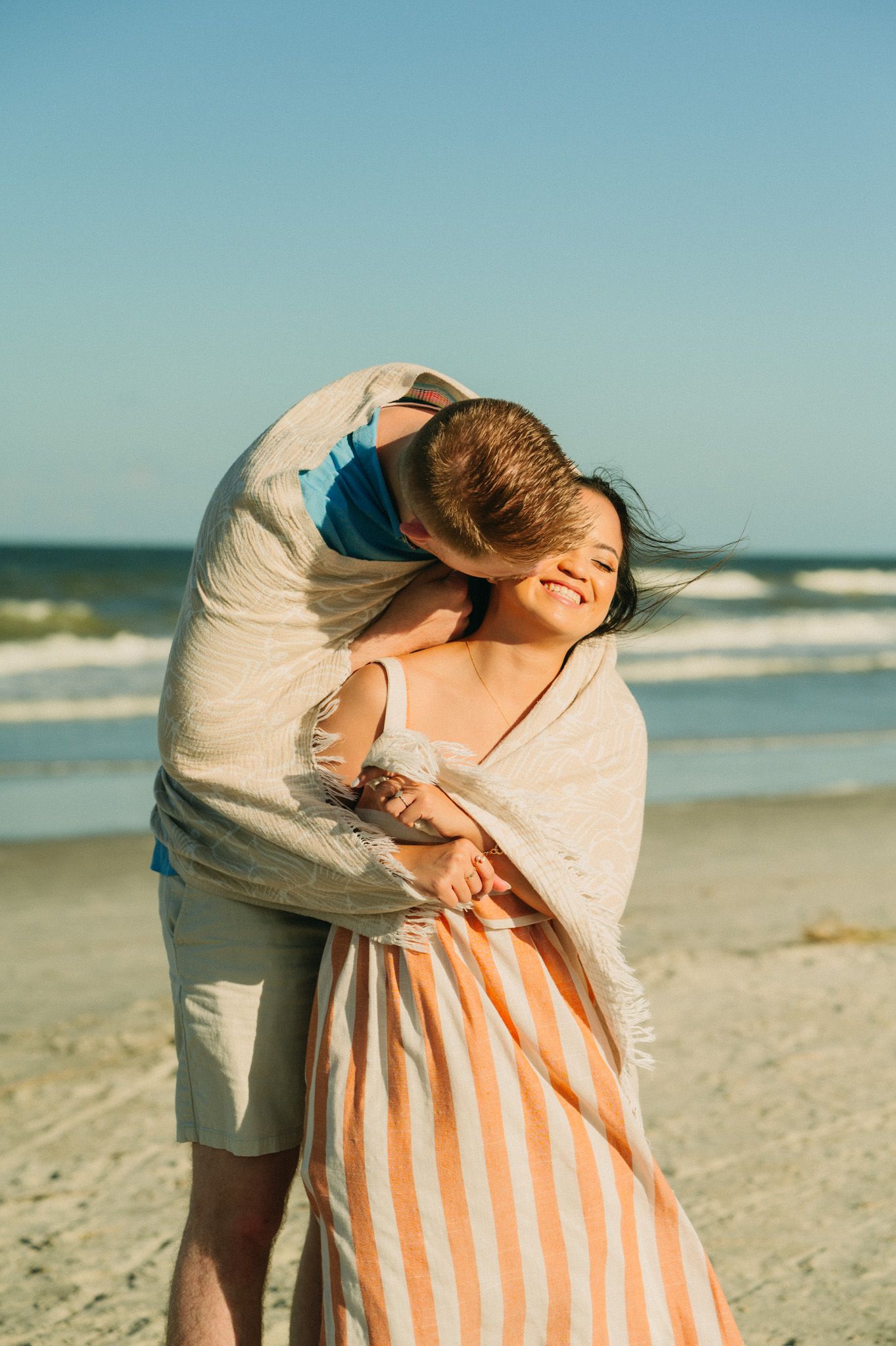 Couple embracing on a sunny beach, woman smiling in striped blanket wrap, waves in background.
