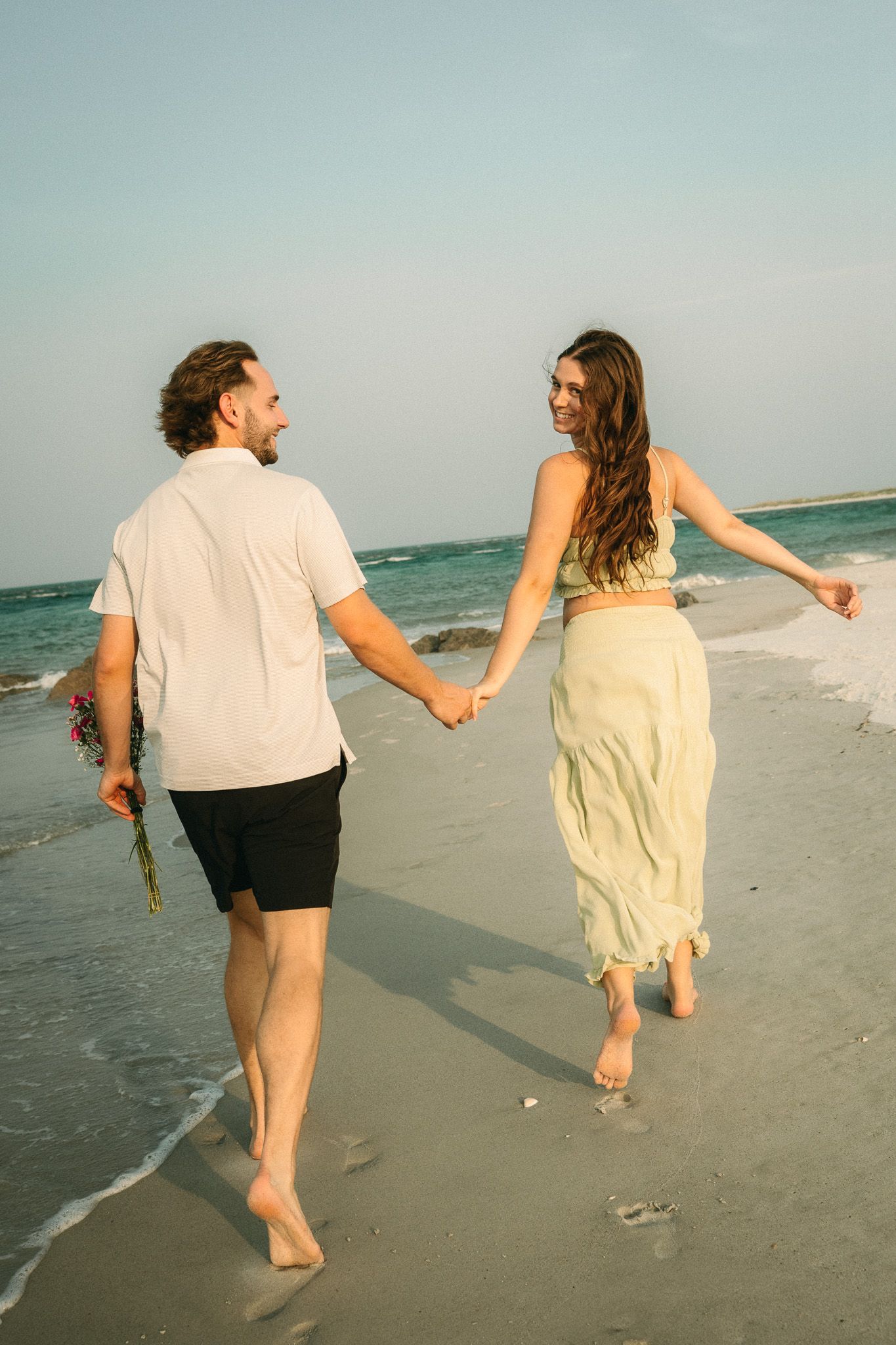 Couple holding hands walking barefoot on beach at sunset, man holding flowers, woman in flowing skirt smiling back.