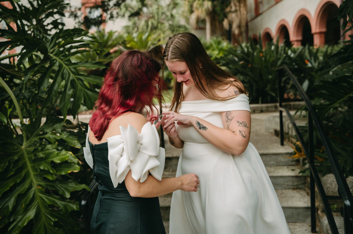 Two brides sharing a tender moment outdoors, one in white off-shoulder gown, other in dark dress, surrounded by tropical greenery.