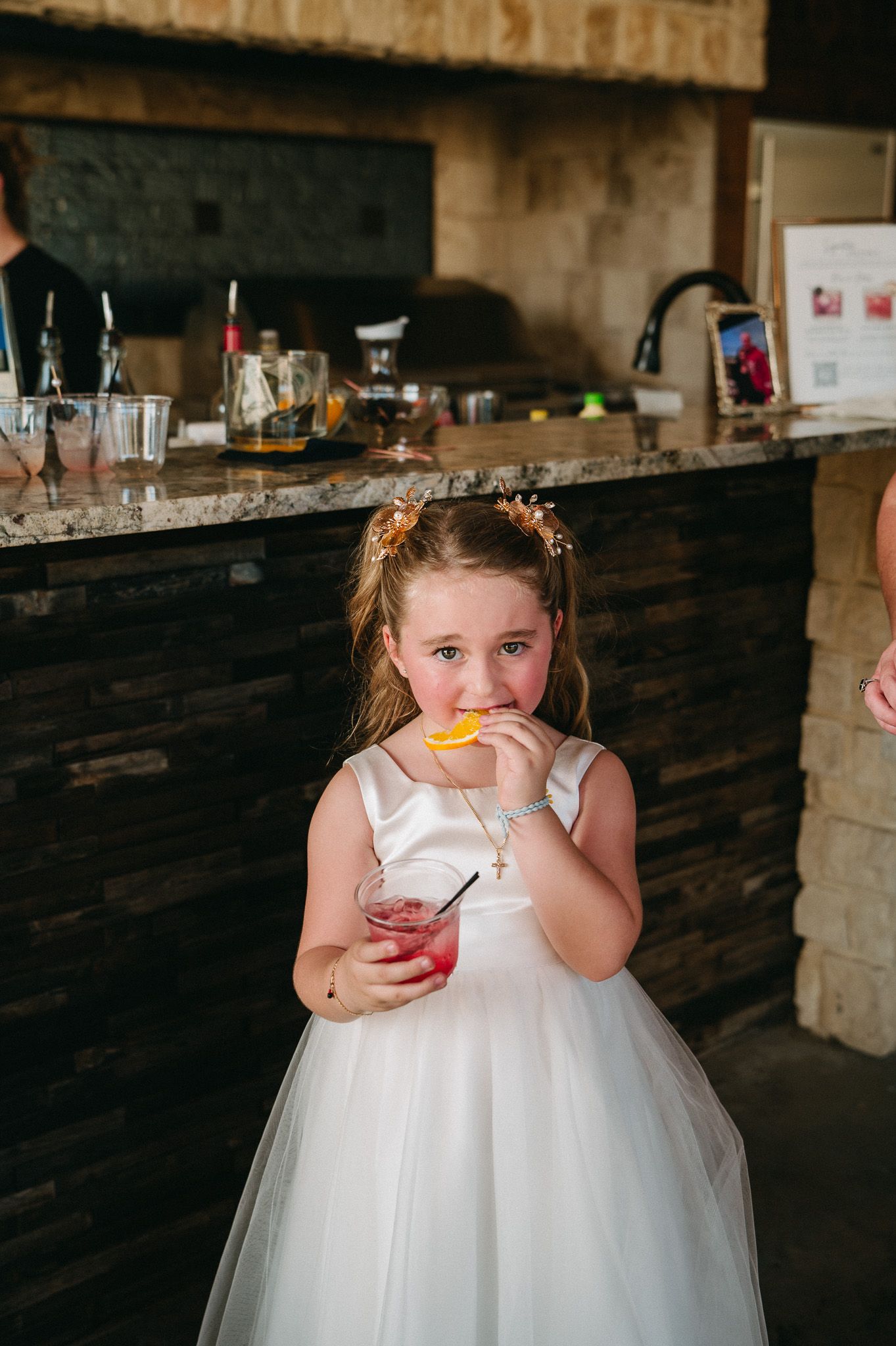 Adorable flower girl in white tulle dress sipping a pink cocktail with orange garnish at wedding bar.