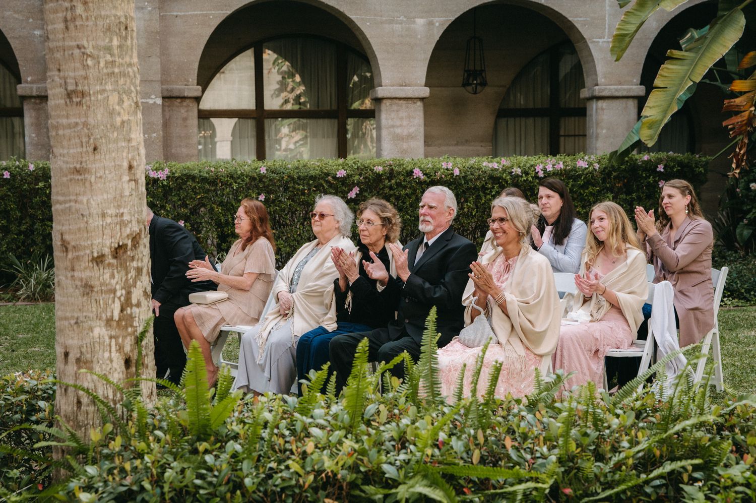Guests in elegant attire clap and celebrate during an outdoor wedding ceremony in a lush garden courtyard.