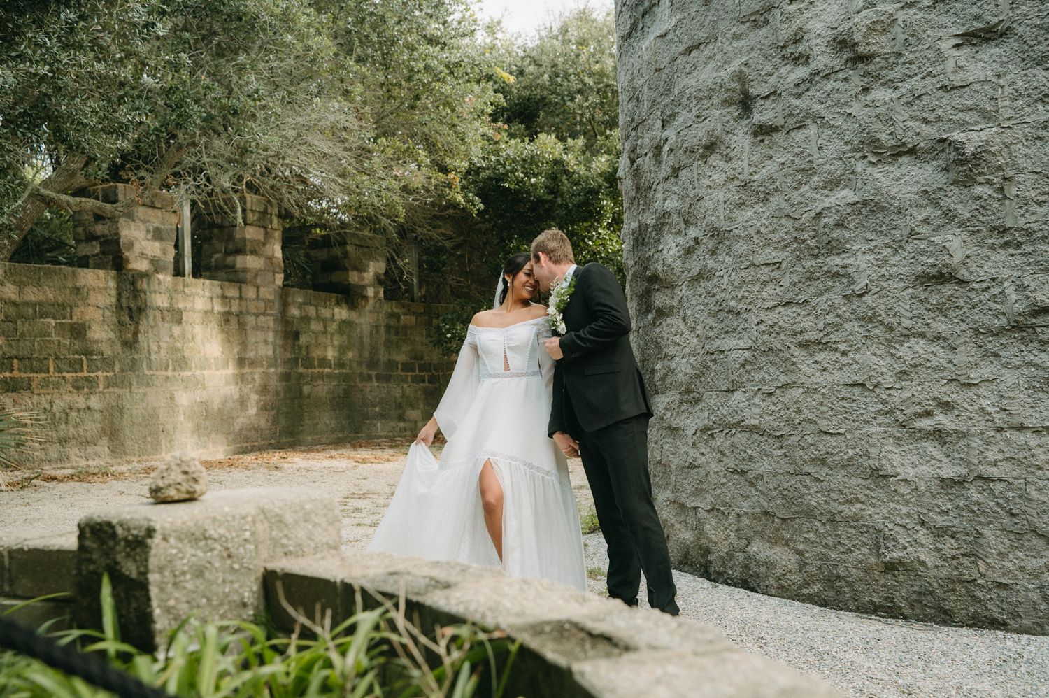 Bride and groom share a romantic kiss outdoors near a stone wall, bride wearing an off-shoulder white gown.