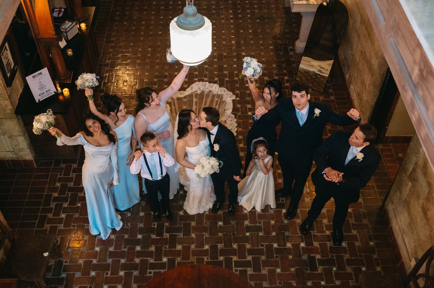 Aerial view of wedding party celebrating with raised arms in historic tiled hotel lobby.
