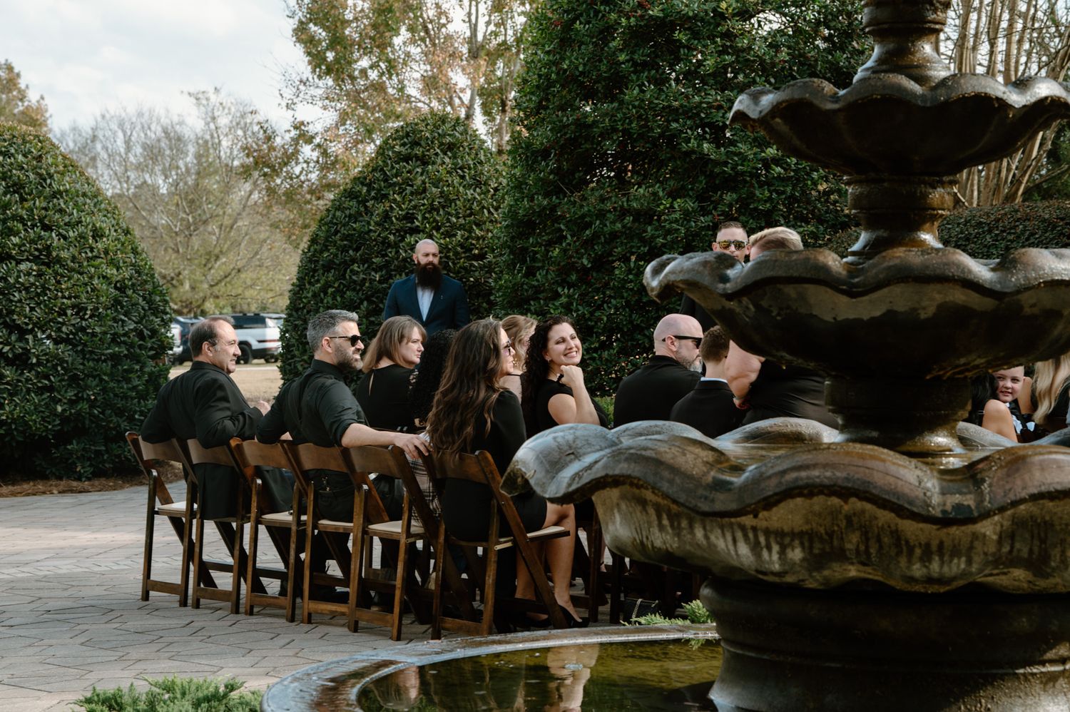 Wedding guests seated outdoors near ornate fountain surrounded by lush green hedges during garden ceremony.
