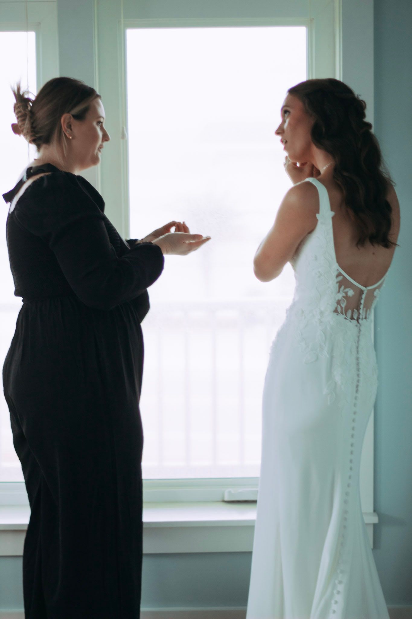 Bride in backless lace wedding gown talking with bridesmaid in black dress by window.
