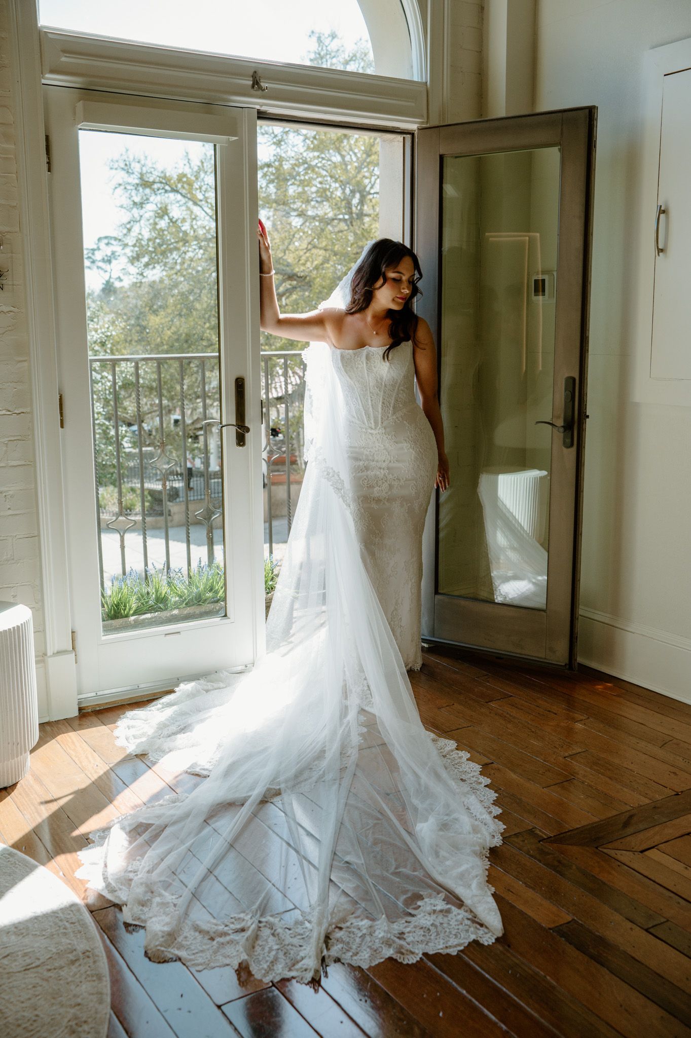 Bride in elegant lace mermaid wedding gown with long train and veil poses by French doors in sunlit room.