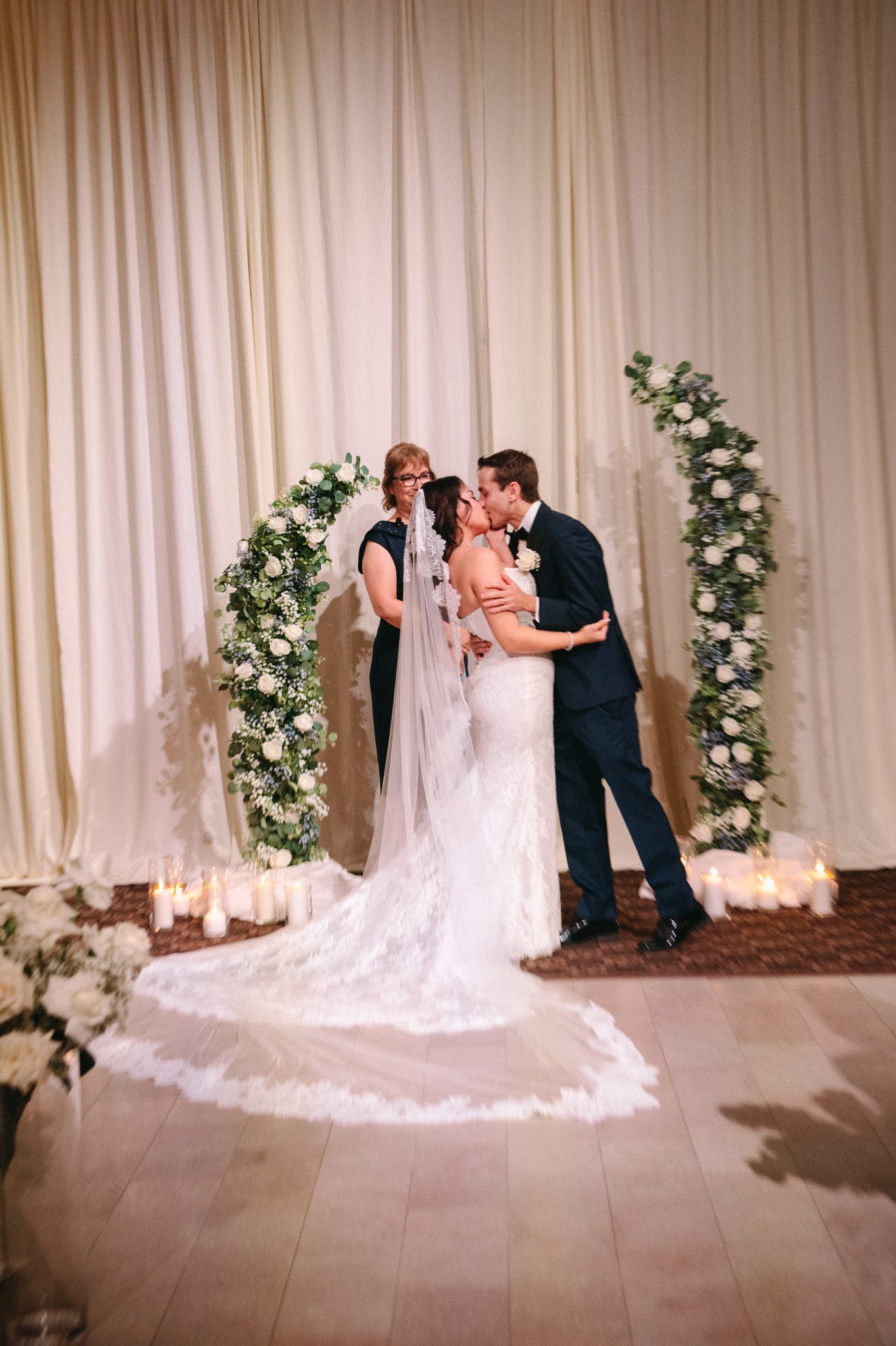 Bride and groom share first kiss at altar beneath floral arch surrounded by candles and white draping.