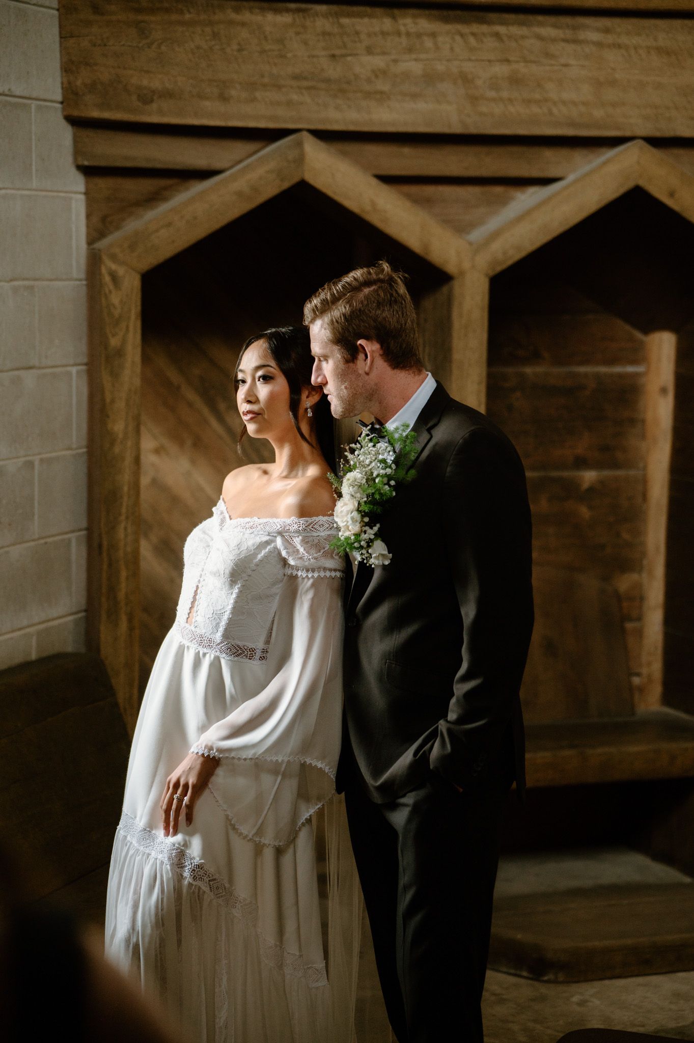 Bride in off-shoulder white gown and groom in black tuxedo pose before wooden geometric backdrop.