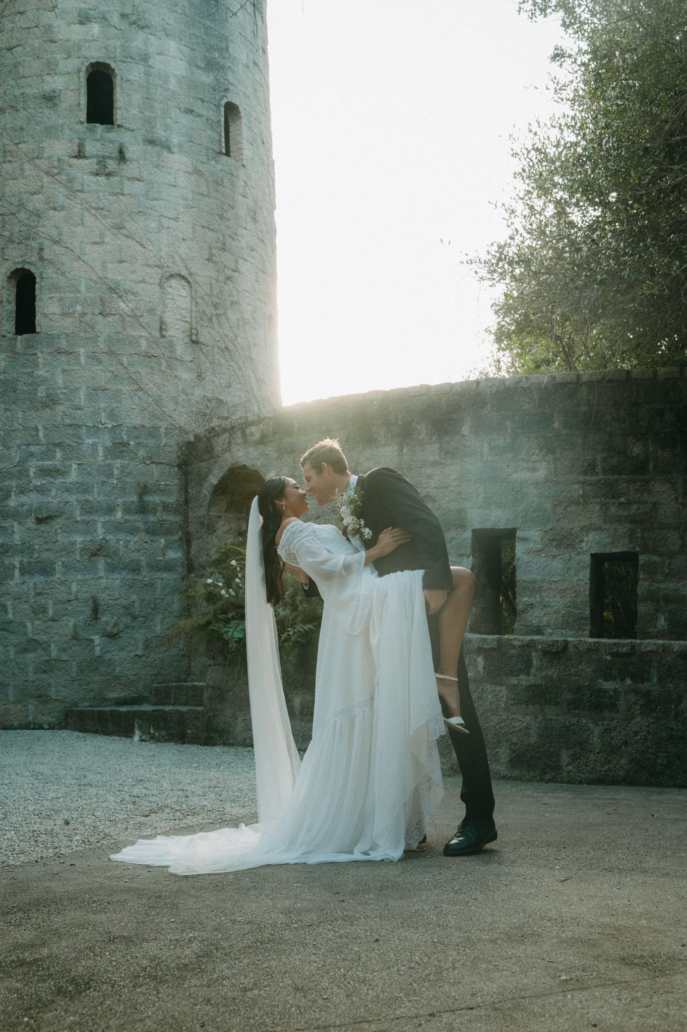 Bride and groom share a romantic kiss outside a sunlit stone castle with flowing white veil.