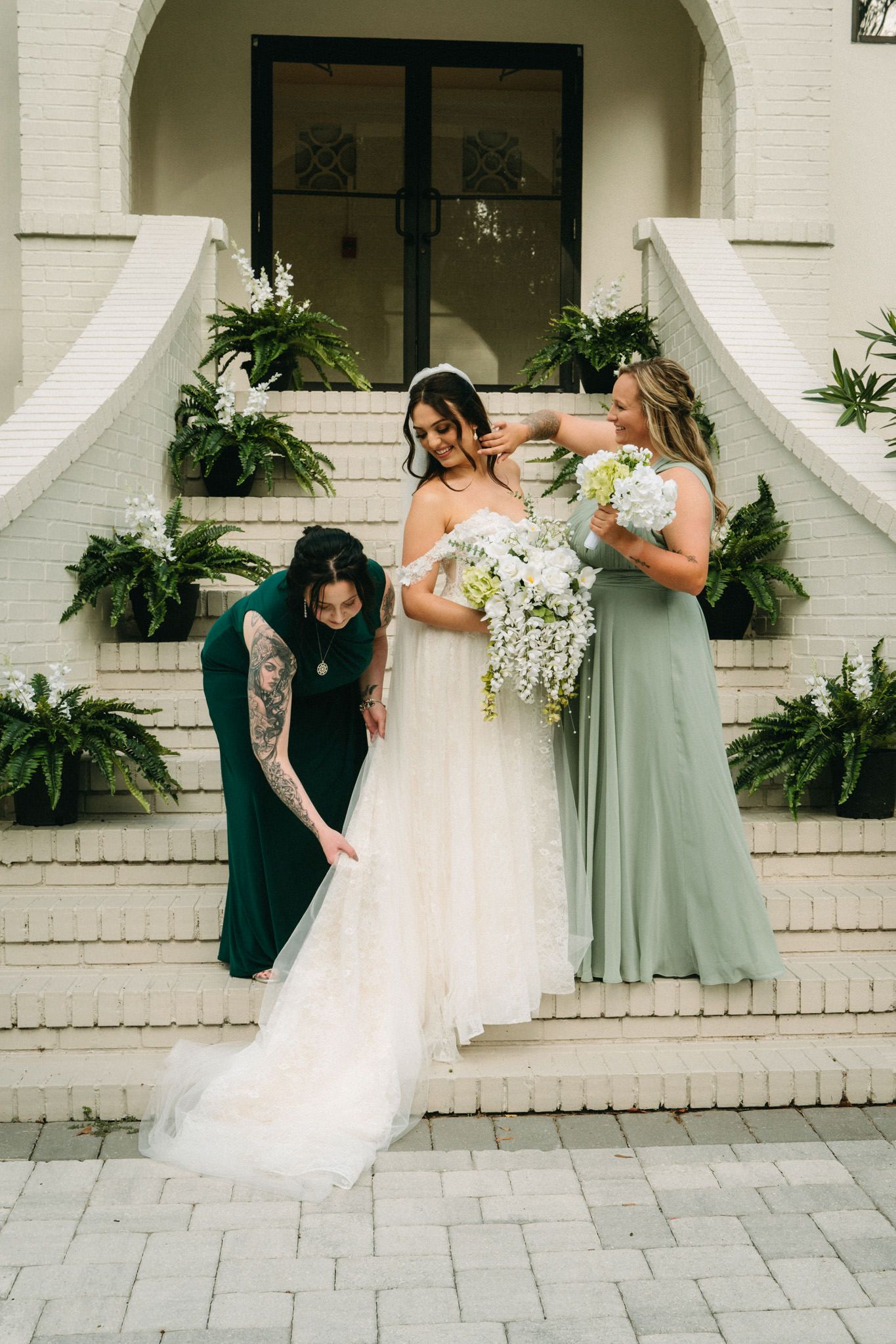 Bridesmaids in green dresses help bride with gown on white staircase.