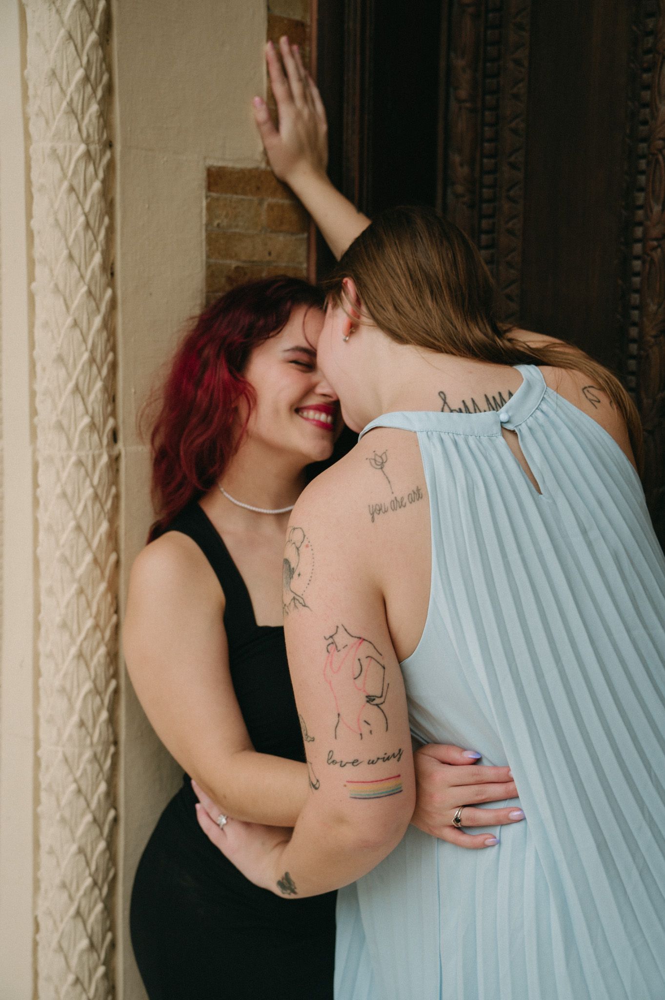 Two women with tattoos laughing and embracing in doorway, one in black dress, one in blue pleated gown.