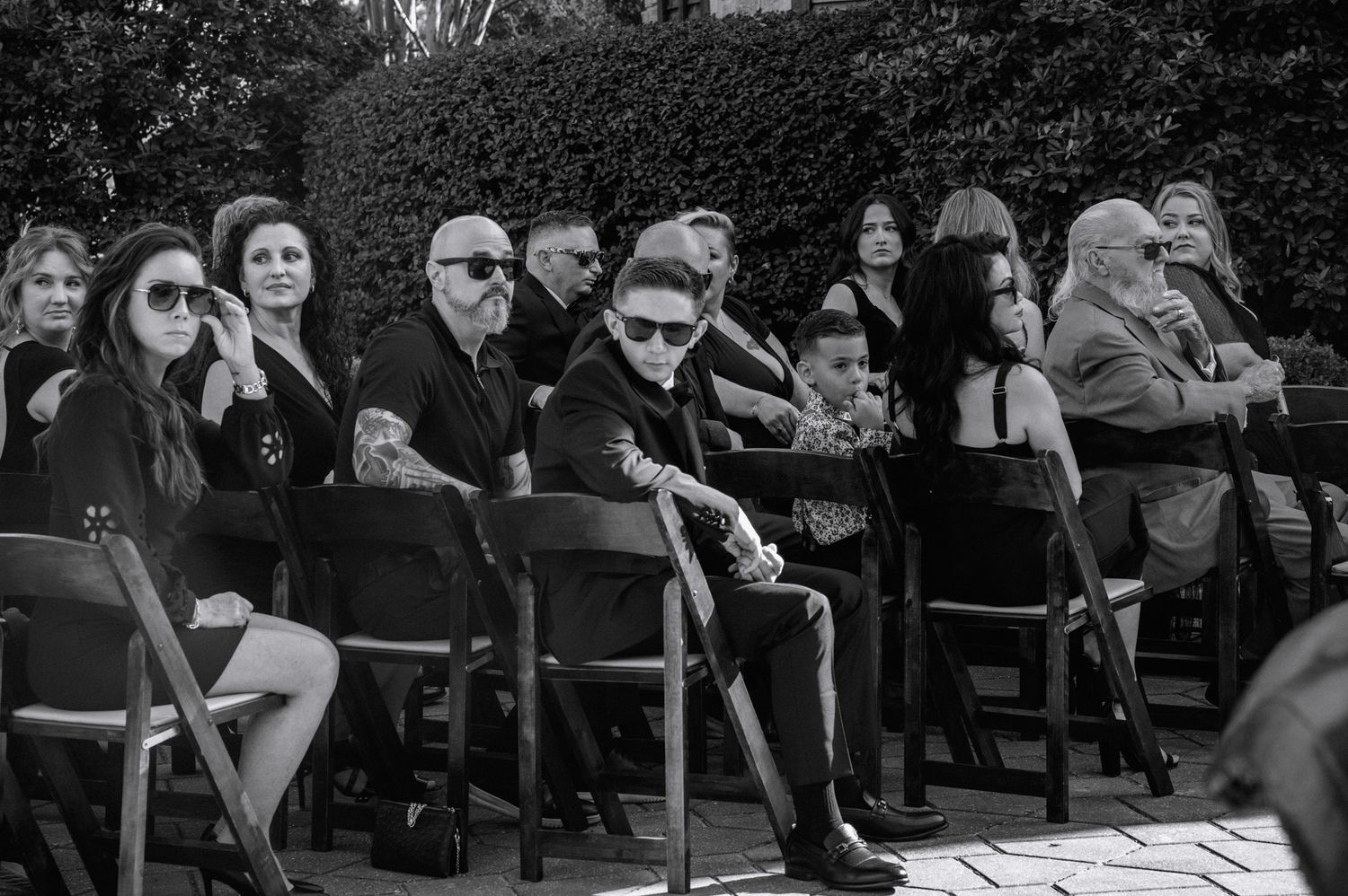 Black and white photo of wedding ceremony guests seated outdoors in front of a lush green hedge.
