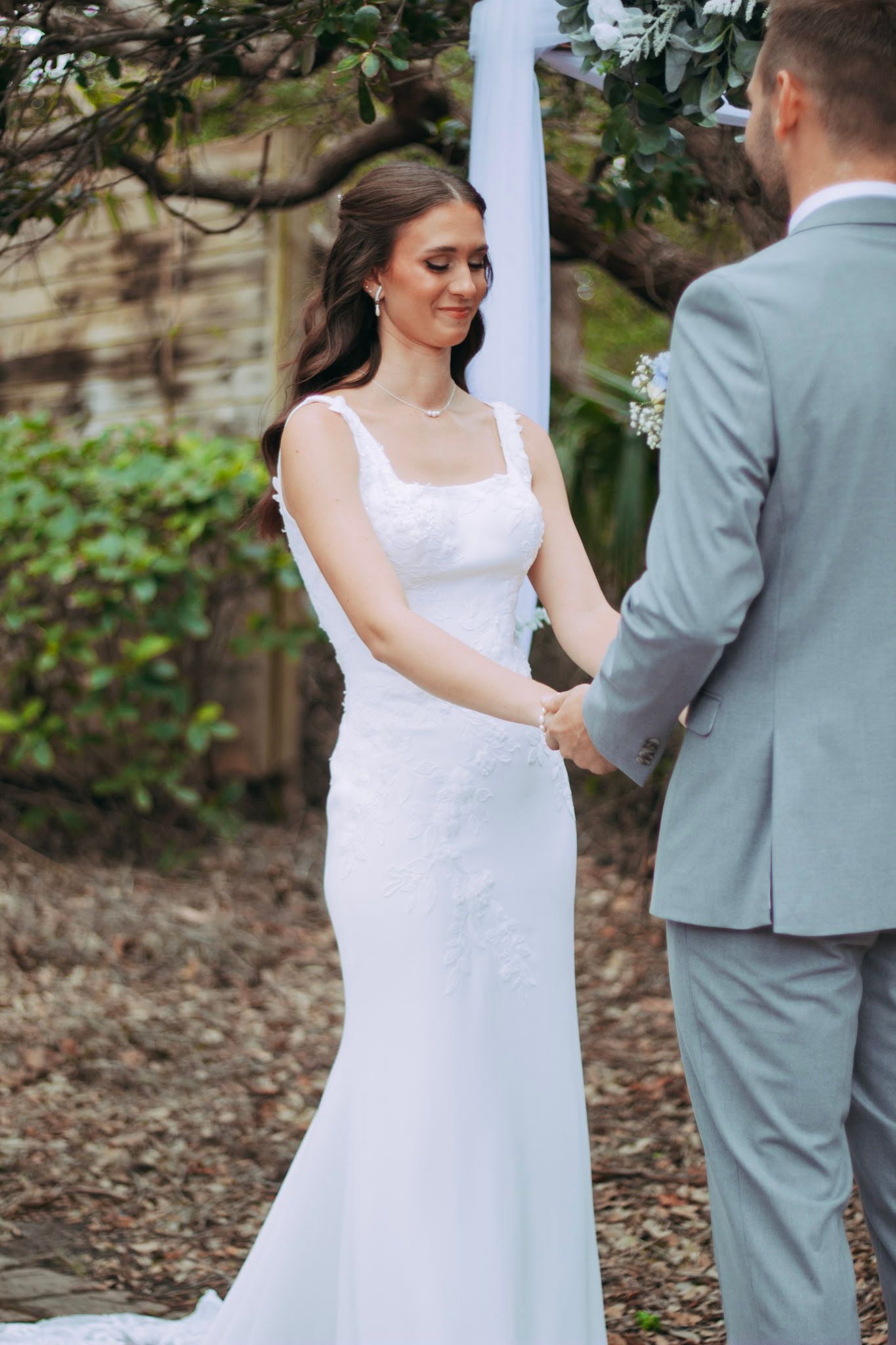 Bride in white lace gown smiles while holding grooms hands during outdoor wedding ceremony.