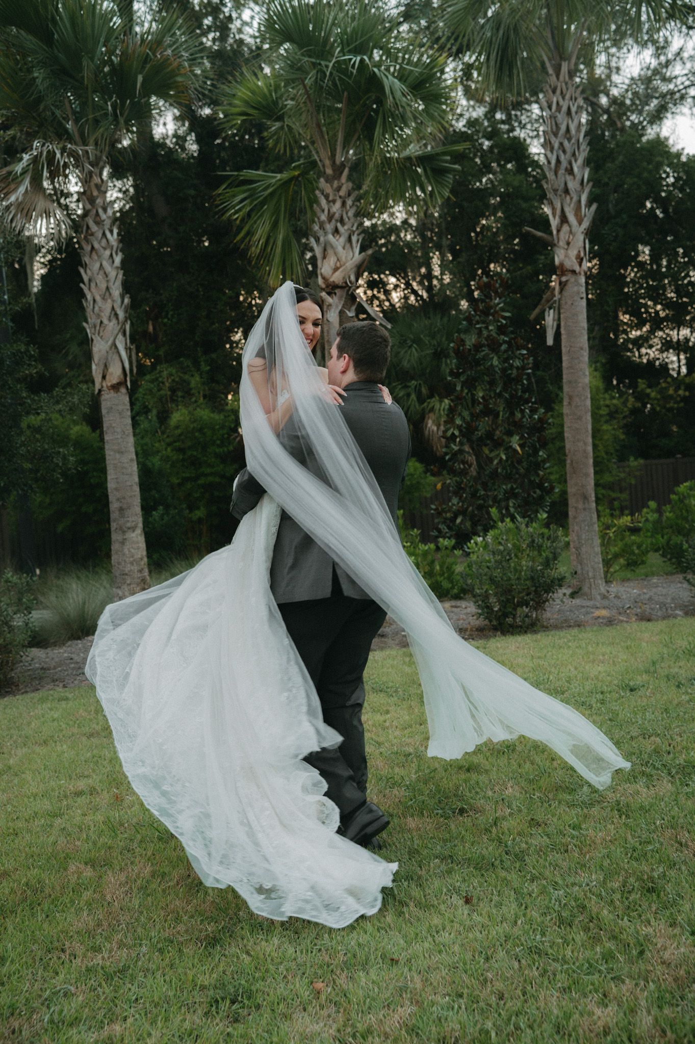 Groom lifting bride in flowing white gown and veil outdoors among palm trees on lush green lawn.