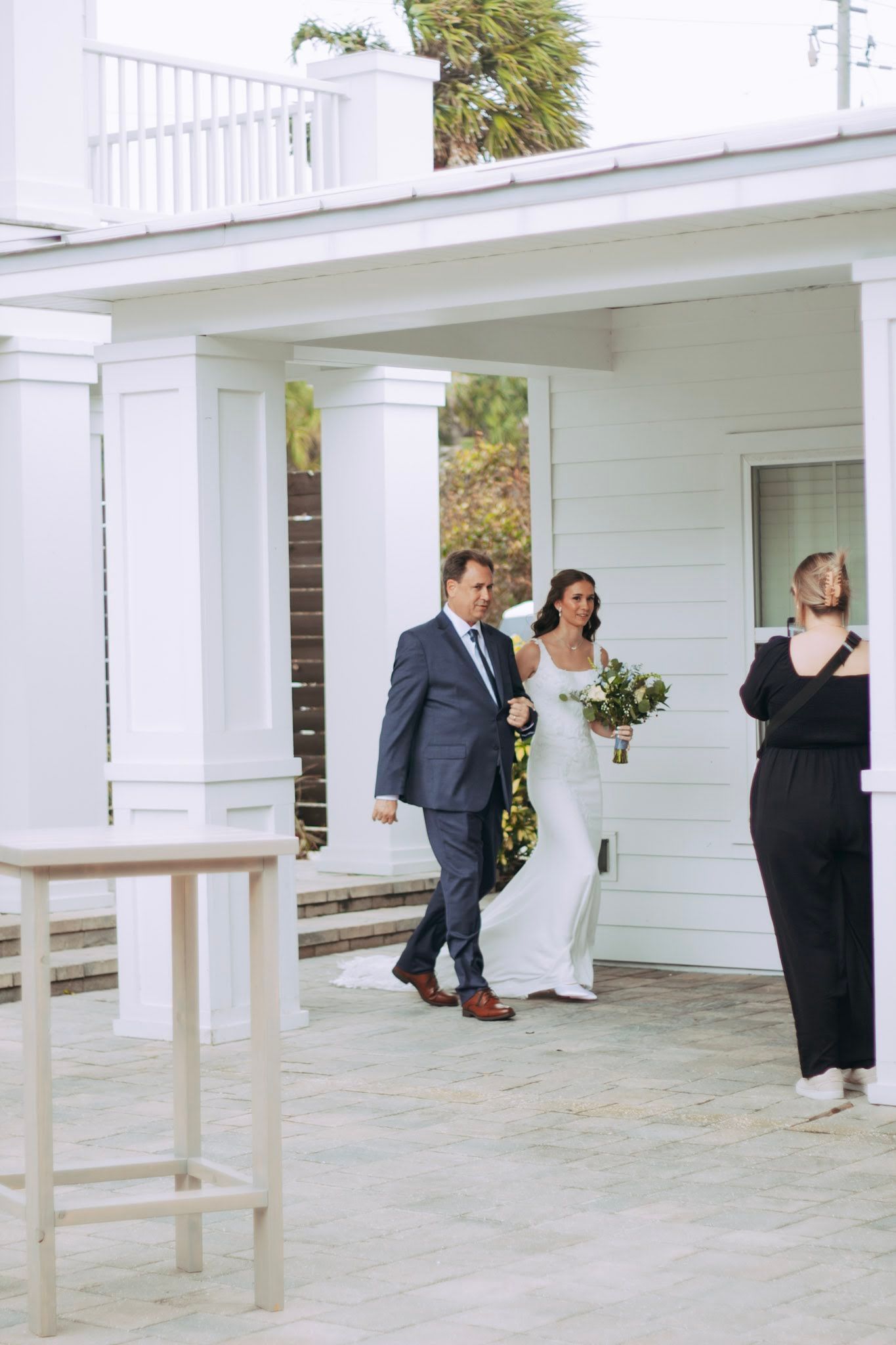 Bride in white gown walks with escort toward outdoor wedding ceremony at white colonial venue.