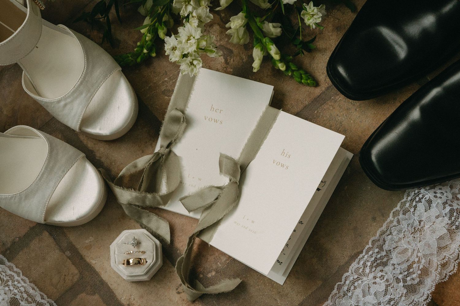 Wedding flat lay with white bridal shoes, black dress shoes, rings, floral bouquet, and elegant ivory vow books with ribbon.