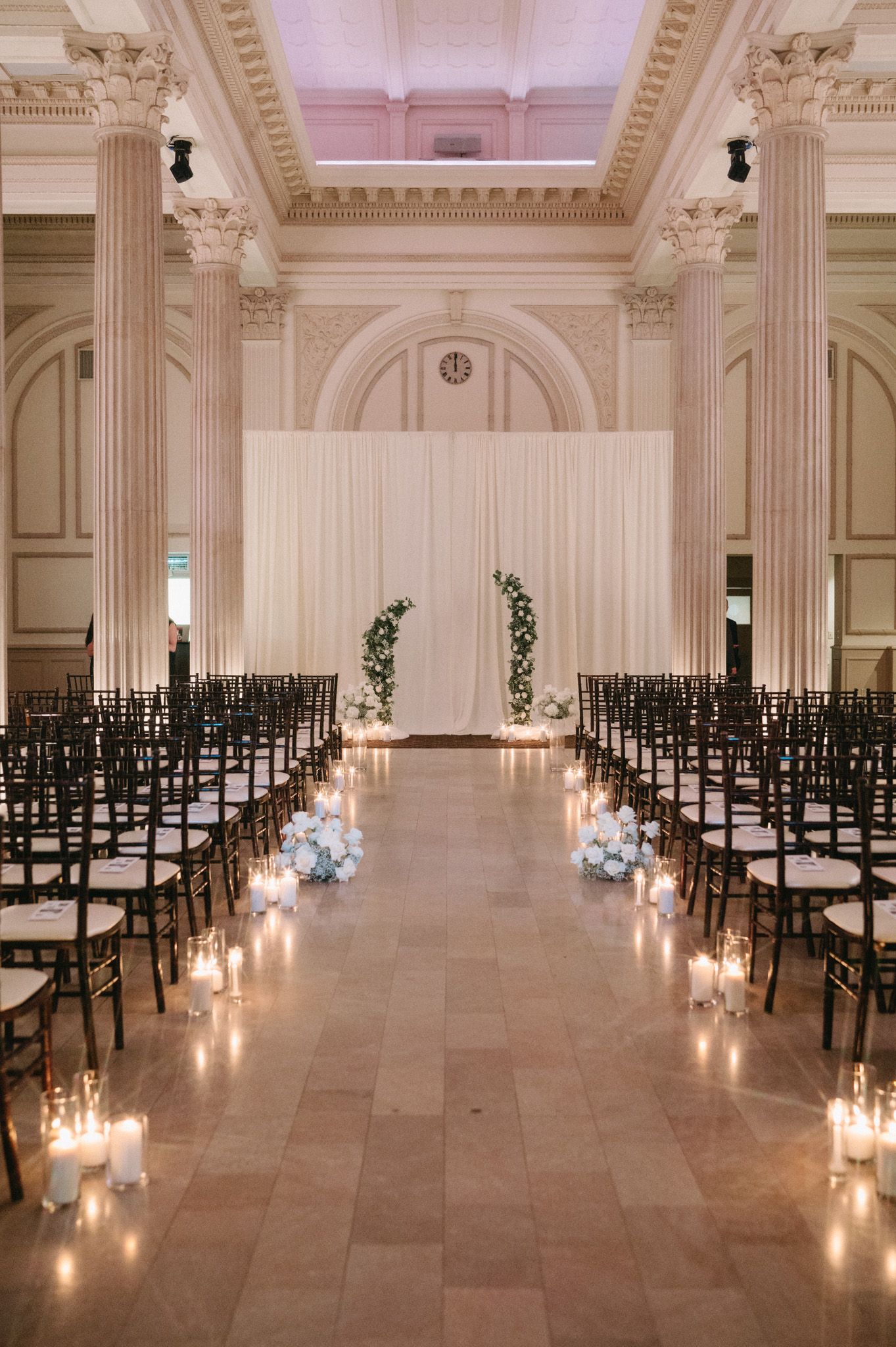 Elegant wedding ceremony setup with candlelit aisle, floral arch, and tall columns in grand ballroom.