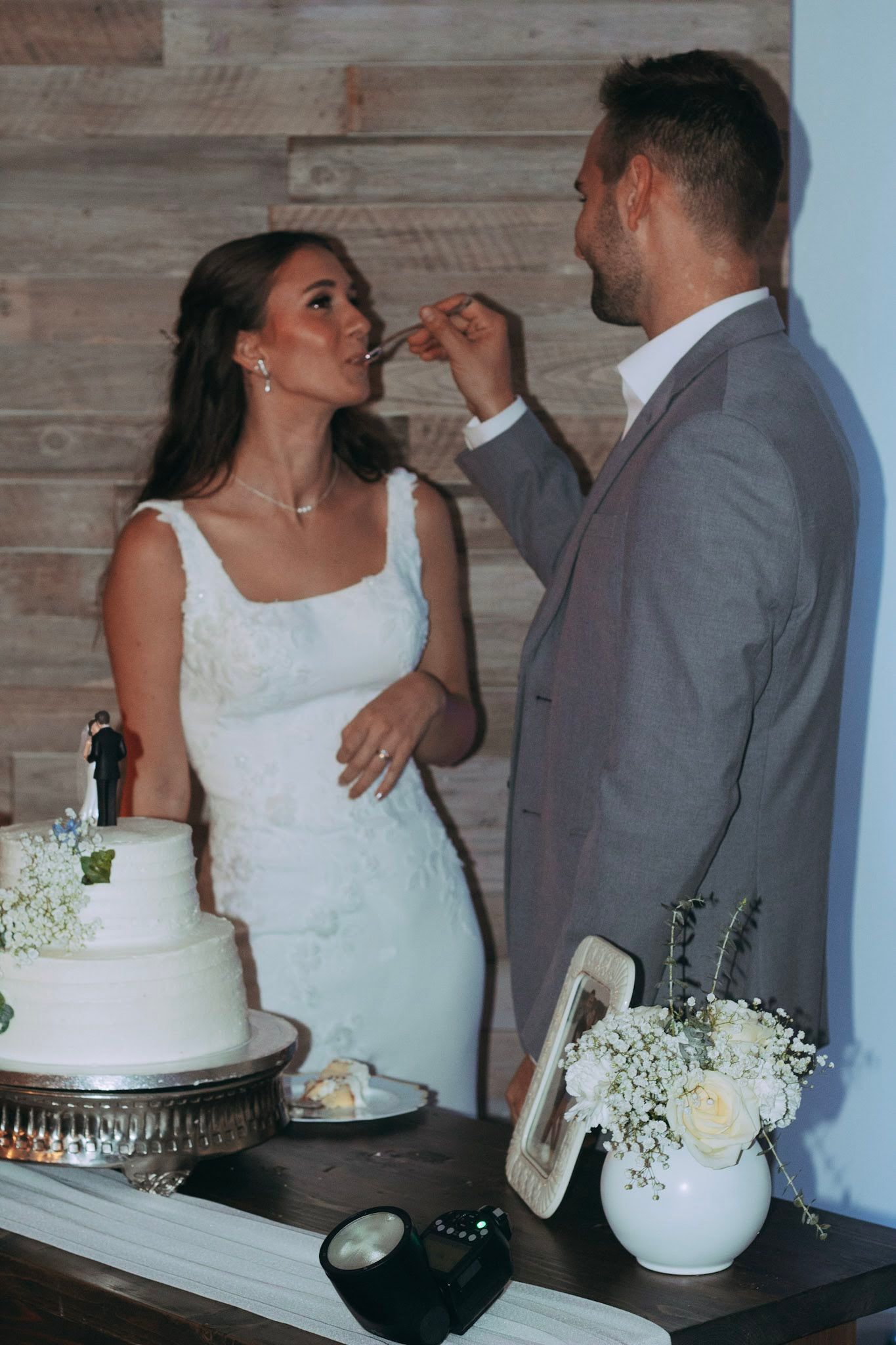 Groom in gray suit feeding wedding cake to bride in white dress at reception table with floral arrangements.